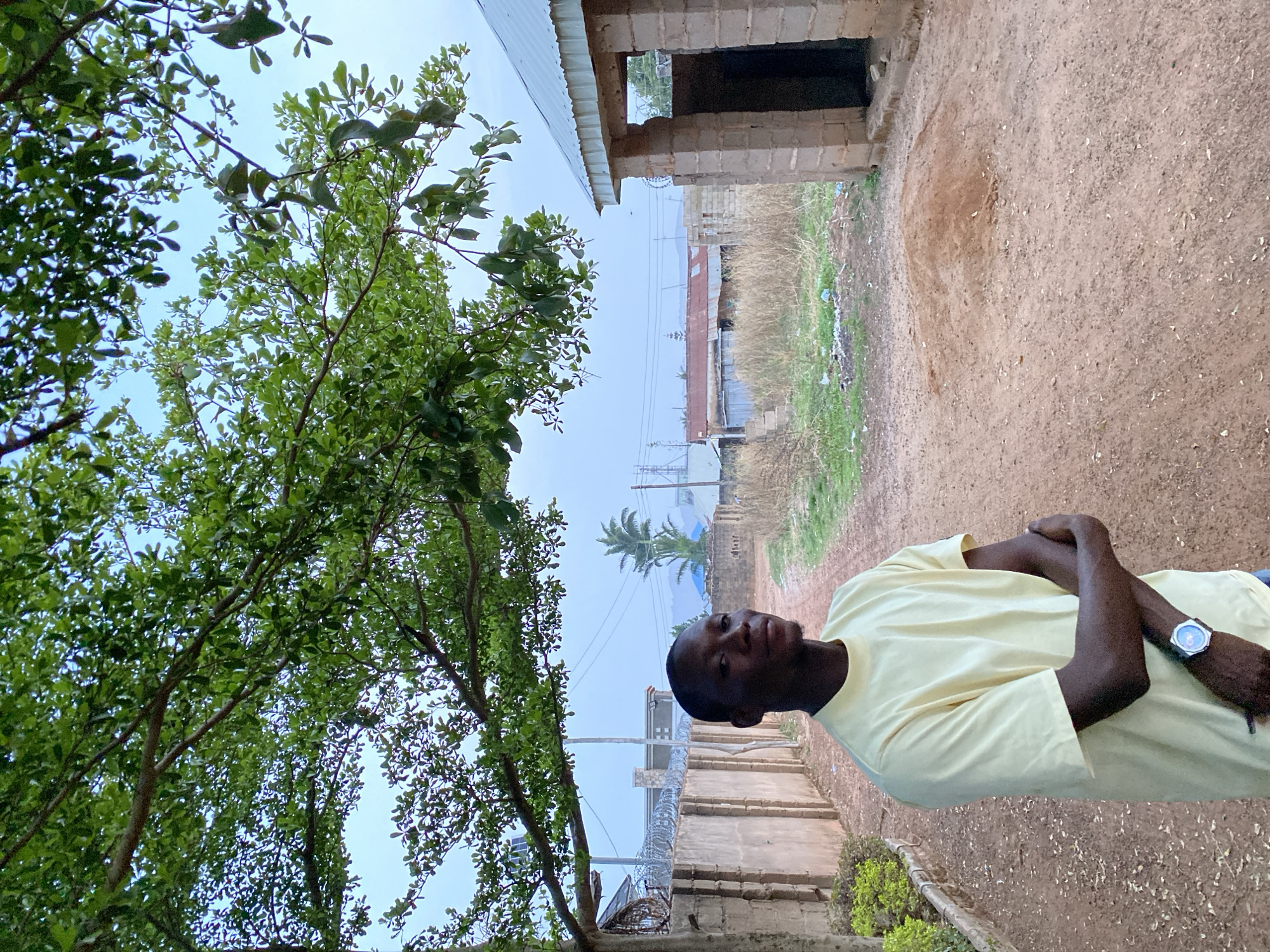 A medium shot reveals a young Black man with short hair, wearing a light yellow t-shirt and a watch on his left wrist. He stands with his arms crossed, facing the camera with a neutral expression.
The setting appears to be a somewhat undeveloped area, likely in Bukuru, Nigeria. A dirt path stretches into the background, flanked by low vegetation and sparse green patches. A partially constructed brick building without a roof is visible on the right. A tree with dense green foliage partially frames the left side of the image. Beyond the brick building, glimpses of houses and a palm tree can be seen in the distance, with utility poles and wires visible against a hazy sky. A grey concrete wall topped with barbed wire is also visible along the foreground of the image. The environment indicates a mixed urban-rural landscape, hinting at a developing neighborhood in Bukuru.