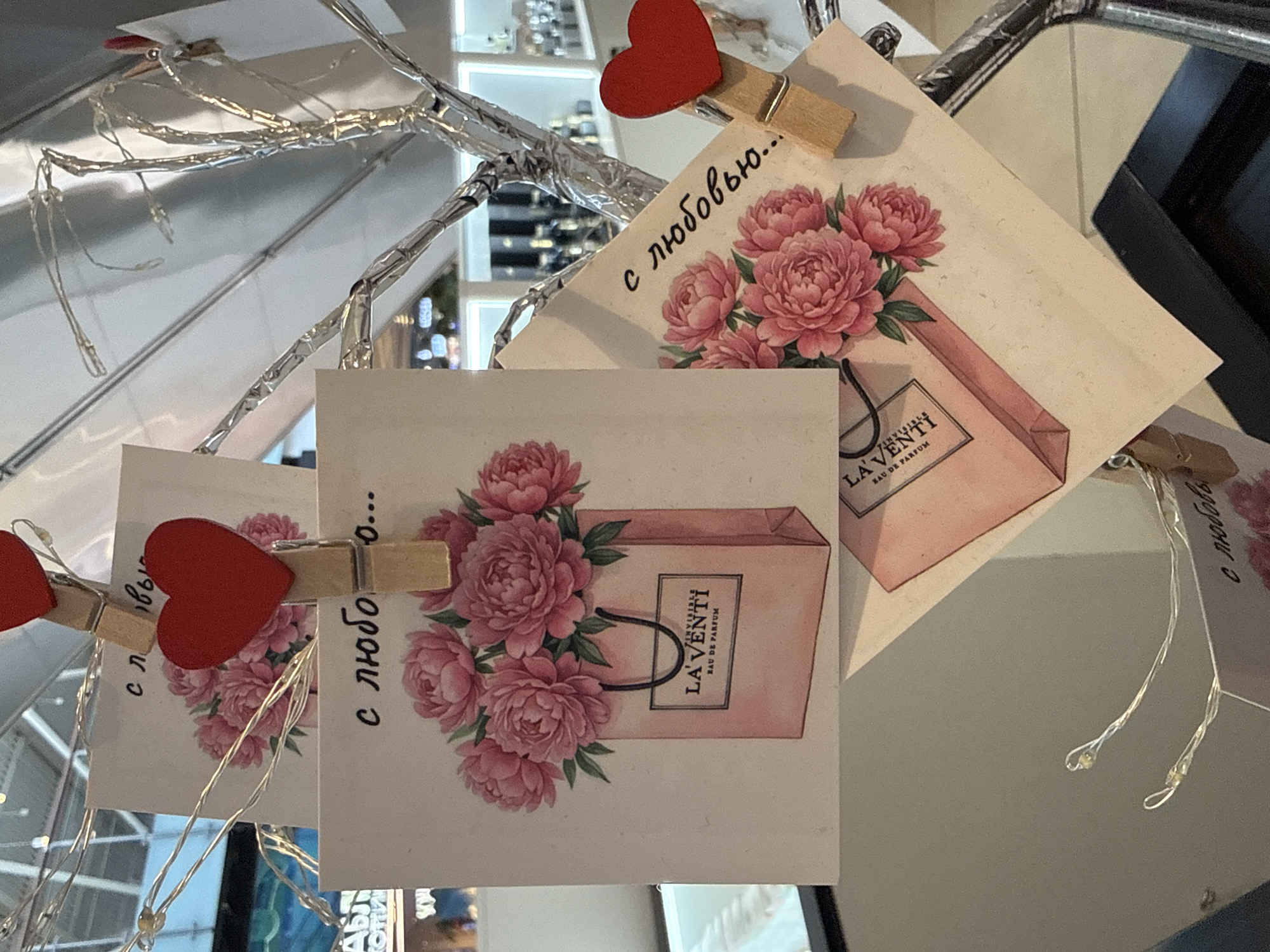 The image shows a close-up of several small cards attached to a decorative, foil-wrapped branch-like structure with clothespins. Each card features a pink peony bouquet and a pink shopping bag with the brand name "LA Venti EAU DE PARFUM" printed on it. Some of the cards have small red heart-shaped decorations. There are also thin, sparkling fairy lights entwined with the branches. The background is blurred, but appears to be an indoor setting with some shelving and reflective surfaces, possibly a store or a decorated display. The text visible on the cards is in Russian and says "с любовью..." which translates to "with love...". One card also has "с любо" partially visible. The overall impression is one of a Valentine's Day or romantic-themed decoration. There are no people visible in the image.