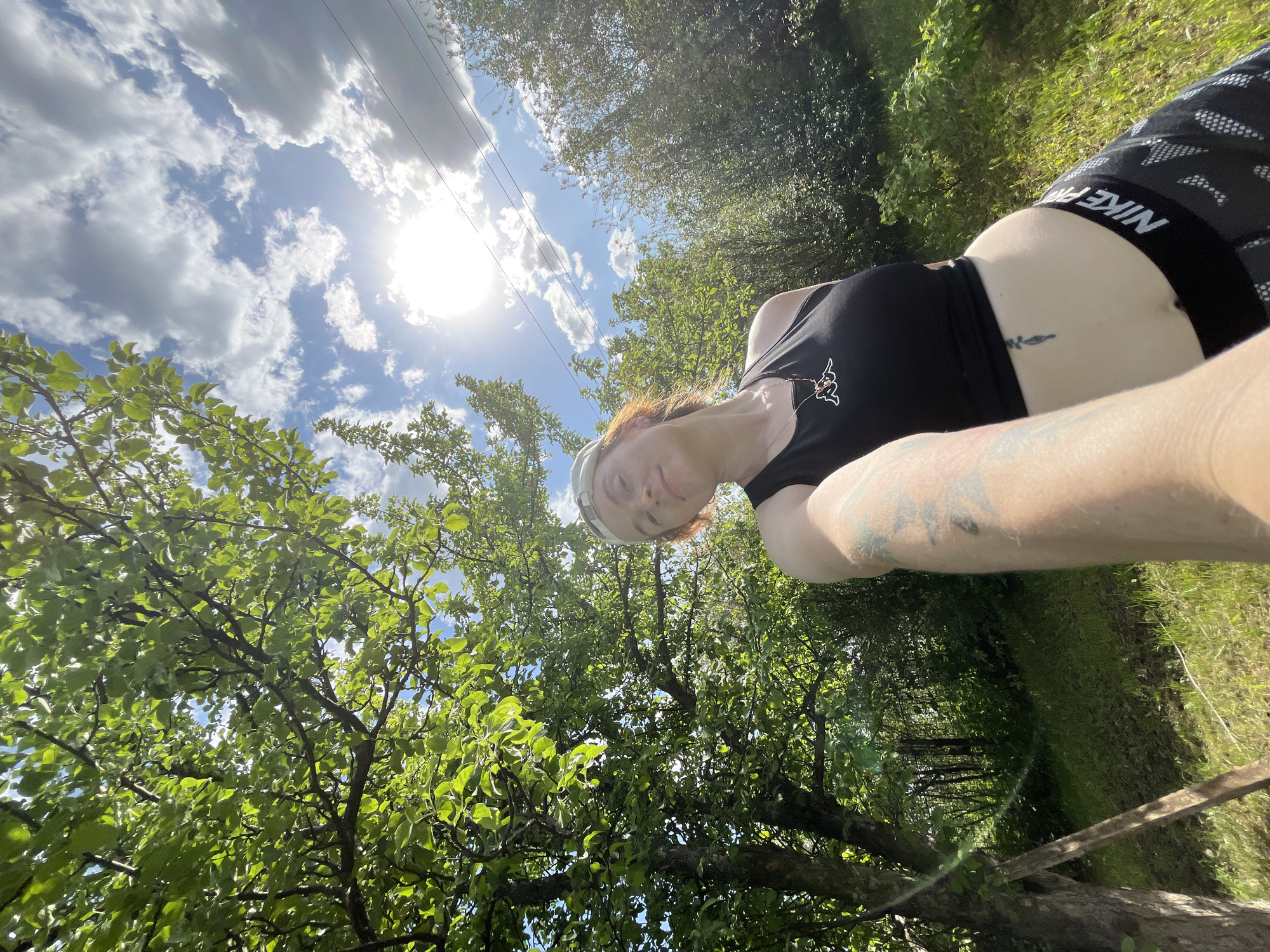 A woman is taking a selfie in an outdoor environment. She is wearing a black tank top with a Kappa logo and Nike branded black and gray patterned shorts. She has visible tattoos on her arm and stomach. The sky is visible in the background, with the sun shining through some clouds. Power lines are also visible in the sky. The environment consists of trees and vegetation, suggesting a natural or park-like setting. This may be in or around the city of Chaikovsky, Russia.