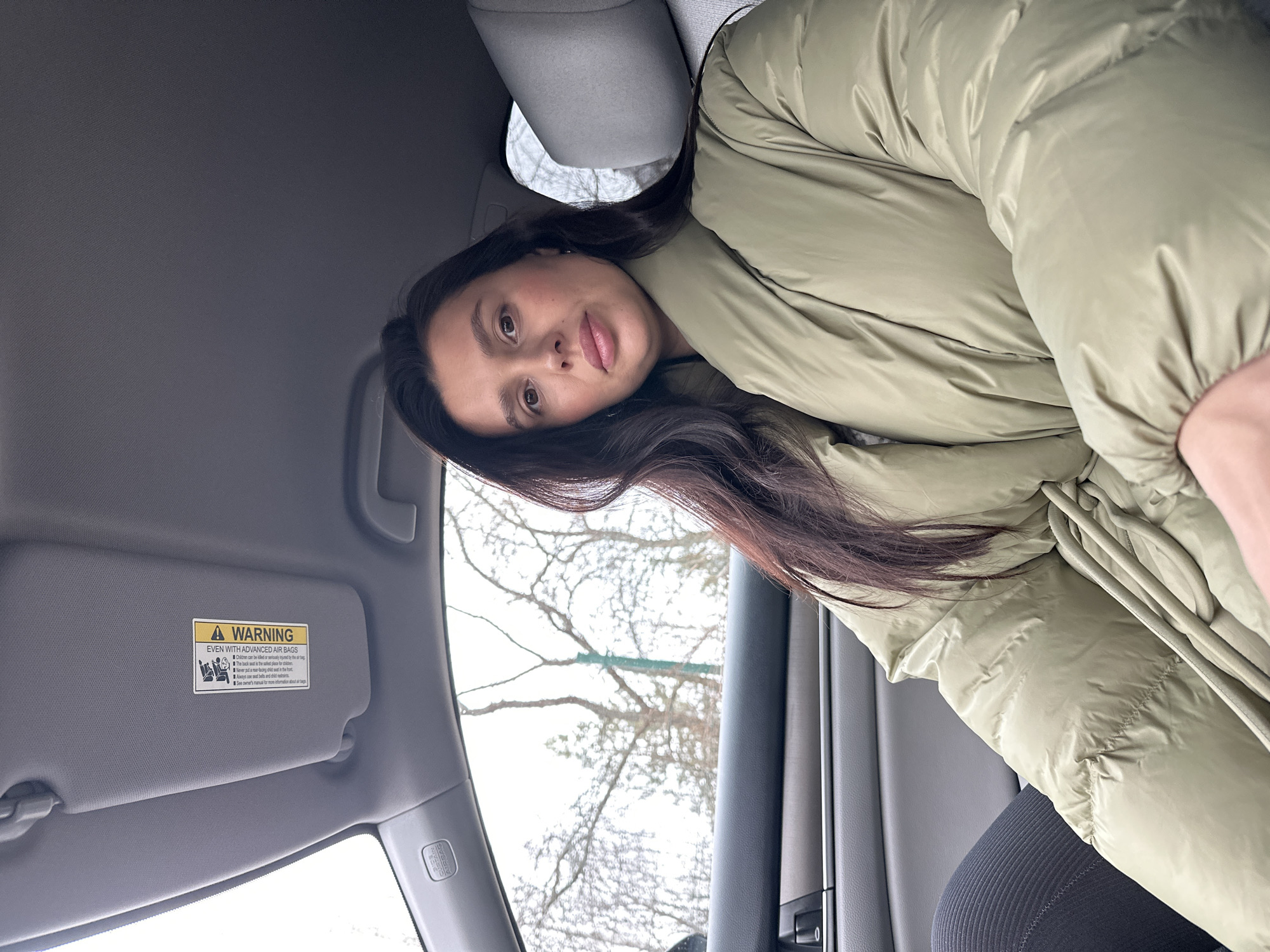 A young woman wearing a green puffy coat is sitting in the driver's seat of a car. The car's sun visor is down, displaying a warning label about airbags. Through the car window, bare tree branches are visible against a gray sky. The location is unknown and no information indicates the city of Chaikovsky, Russia.