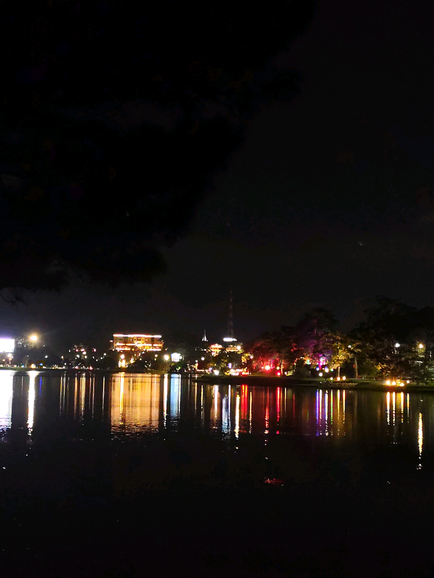 The image depicts a nighttime scene of Xuan Huong Lake in Da Lat, Vietnam, with the cityscape illuminated by artificial lights. The foreground is dominated by the dark, reflective surface of the lake, mirroring the lights of the city and surrounding trees. In the mid-ground, a large, brightly lit building with multiple stories and prominent windows stands out, emitting a warm, orange glow. To the right of this building, several trees are illuminated with colorful lights, creating a vibrant and festive atmosphere. A tall, slender tower, possibly a communication tower, is visible in the background. The sky is dark and cloudy, with a few faint stars or distant lights. The overall impression is of a tranquil, picturesque urban landscape at night, with the lake serving as a central element reflecting the urban ambiance. There are no visible people or specific actions taking place in the image.