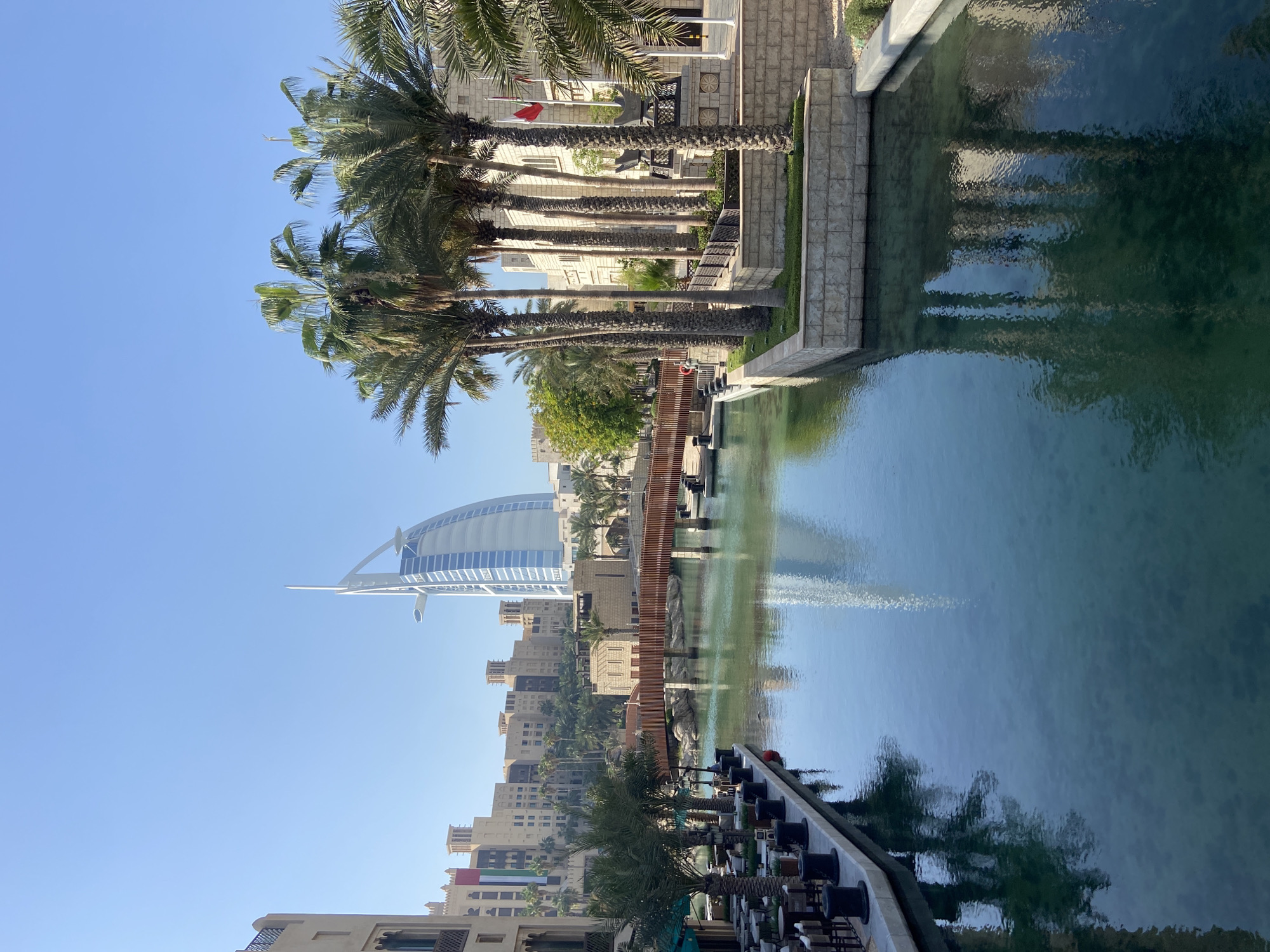 The image shows a view in Dubai, United Arab Emirates, with the Burj Al Arab hotel prominently visible in the background. A body of water, possibly a canal or artificial lake, dominates the foreground, reflecting the buildings and sky. Palm trees line the edge of the waterway on both sides. The architecture in the midground includes traditional Middle Eastern-style buildings with a distinctive sand-colored facade. A wooden walkway spans the water. The sky is clear and blue.