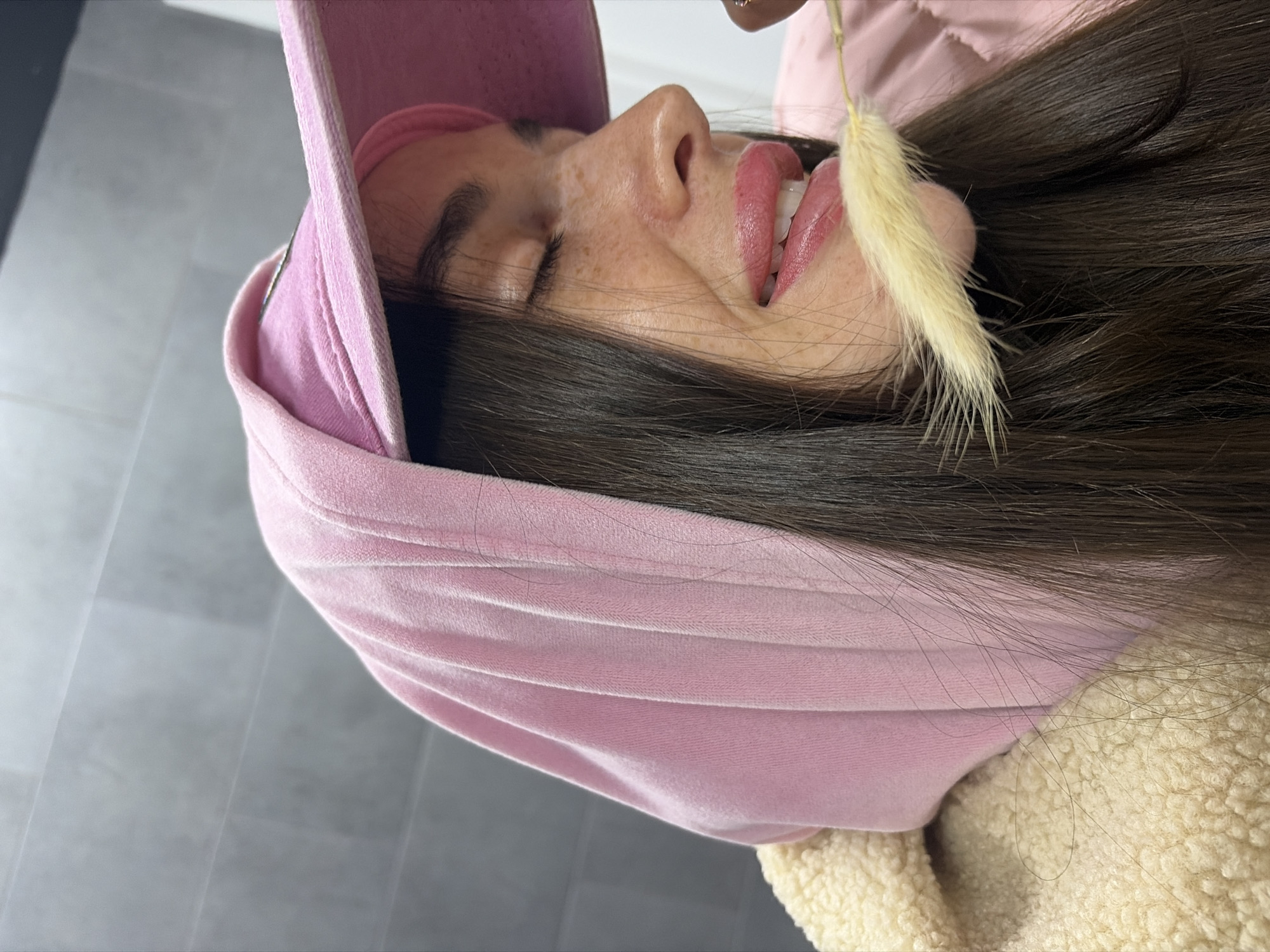 A side-view indoor shot depicts a woman with brown hair, freckles, and pink lipstick wearing a pink cap, with a decorative plant stem near her face. The setting appears to be a room with a grey tiled floor. The location, Saint Petersburg, Russia, is not ascertainable from the visual information provided.