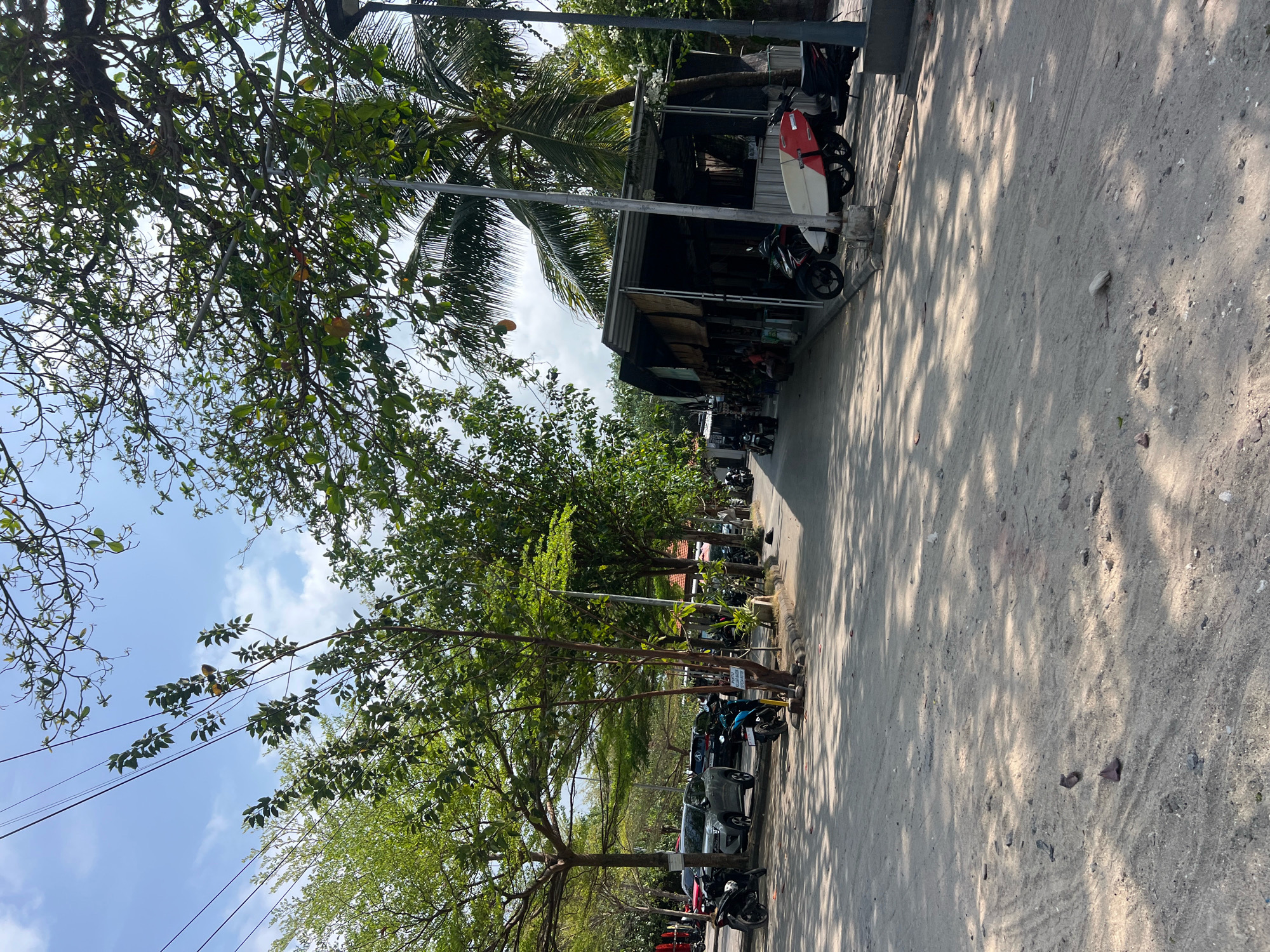 A dusty, sun-drenched street in Kuta, Indonesia, is lined with parked motorbikes and a few cars. Large trees with full green canopies cast dappled shadows across the sandy ground. In the background, a small, open-air structure, possibly a shop or cafe, is visible, with more motorbikes parked in front. A red and white surfboard leans against the structure. The sky is a bright blue with a few wispy clouds, and power lines are strung between trees. The scene appears to be a typical streetscape in a tropical, coastal area.