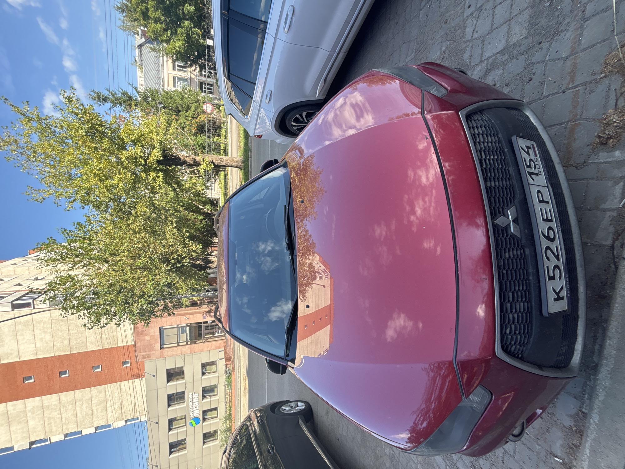 A red Mitsubishi Lancer, license plate K526EP 154 RUS, is parked on a cobblestone street in front of a modern building with a red and beige facade. The building has several windows and a sign that reads "БАЛТИЙСКИЙ ЛИЗИНГ" (Baltiyskiy Lizing) in Cyrillic script, indicating a financial leasing company. To the right of the Lancer is a silver SUV. Trees with green foliage are visible in the background, along with a clear blue sky with some clouds. The image appears to have been taken on a sunny day in Novosibirsk, Russia, given the context clues from the license plate and the building's signage.