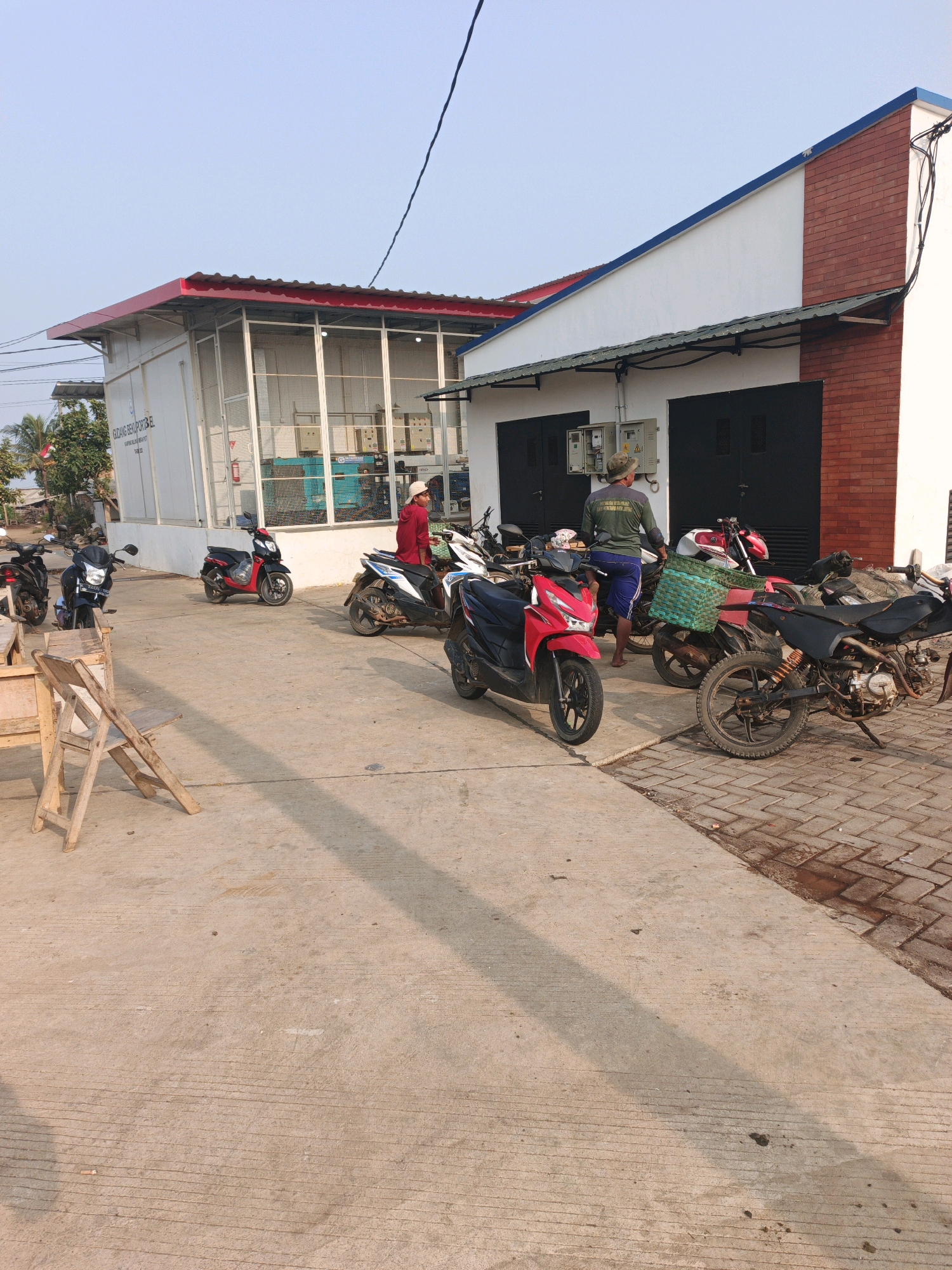 In Bumiharjo, Indonesia, on a paved outdoor area with a concrete and brick building, several motorcycles are parked. Two individuals are visible. One person, wearing a red top and a white cap, stands next to a white and black motorcycle. Another person, wearing a green shirt and blue shorts, is near a motorcycle with a green basket. The building has a white facade with red brick accents and large, dark garage doors. A white structure with a red roof and metal fencing is also part of the scene, with visible machinery inside. A wooden folding chair and table are positioned on the concrete in the foreground. The sky is clear and blue, suggesting daytime.