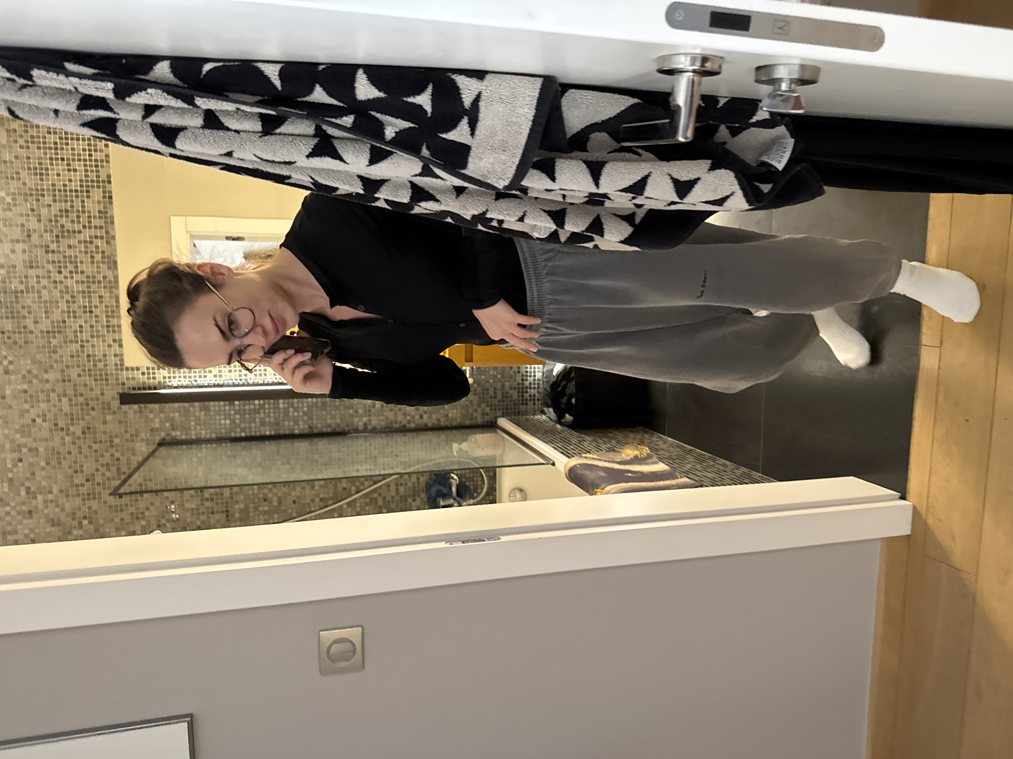 The image, taken indoors in Warsaw, Poland, shows a woman in what appears to be a modern bathroom or an area adjacent to it. The woman, with light brown hair tied back and wearing glasses, is dressed in a black long-sleeved top and grey sweatpants, with white socks. She holds a dark-colored phone or small device near her face, seemingly looking at its screen with a focused, neutral expression.
The setting features a prominent white door frame in the foreground, from which a thick, folded towel with a distinctive black and white geometric triangle pattern is hanging. Visible behind the woman is a shower area with walls covered in small, light brown mosaic tiles. A clear glass partition separates the shower space. Inside the shower, a dark blue and gold patterned item (possibly a bath sponge or loofah) rests on a shelf. The floor transitions from dark grey tiles in the immediate area to light wood planks further into the room. A silver-colored light switch or electrical outlet is visible on a grey wall segment. Two chrome door stoppers or latches are mounted on the top white door frame. A small tag on the towel is partially visible, showing the text "MONO". The lighting suggests an indoor environment, possibly with artificial light.