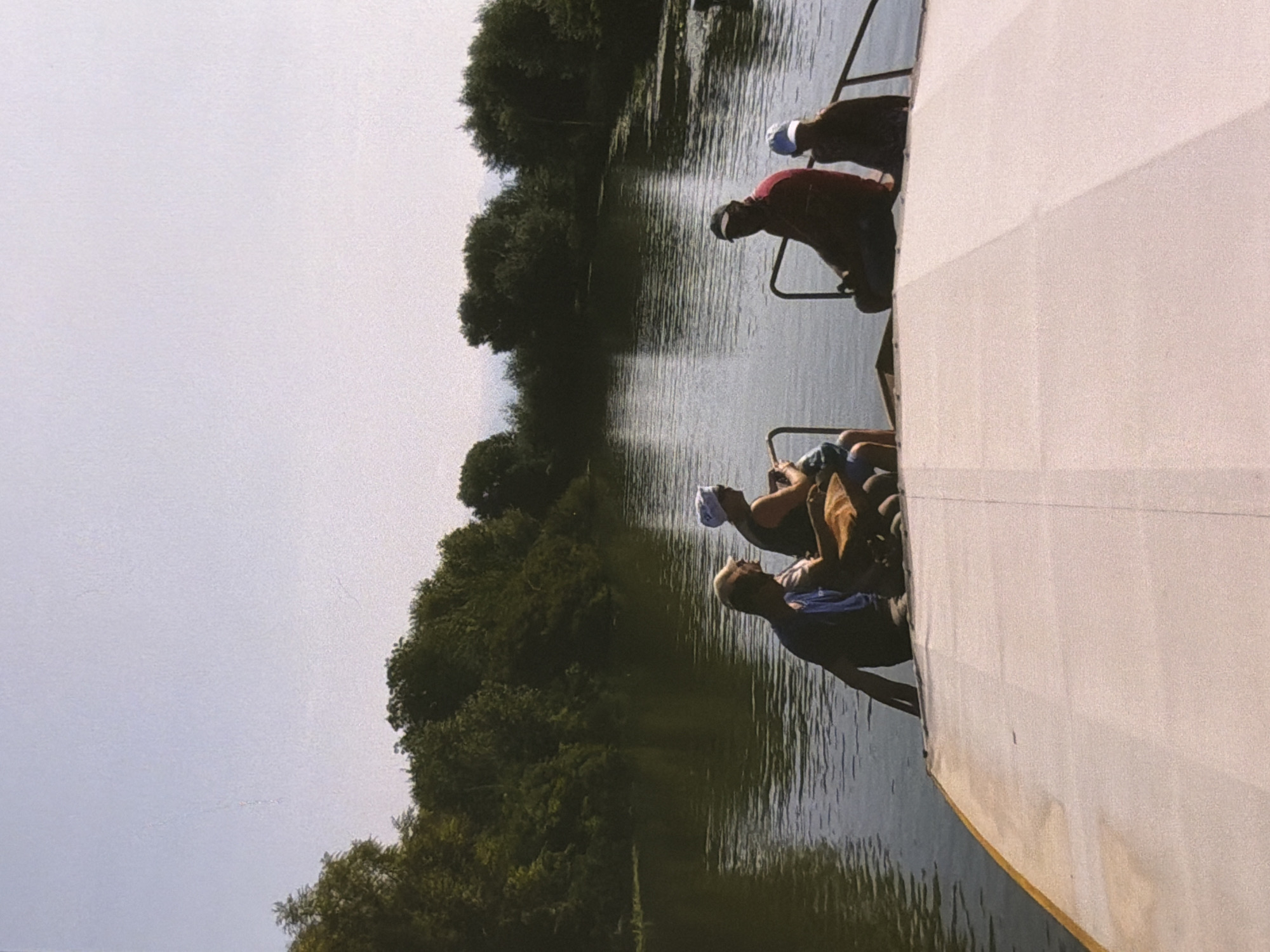 A group of five people are sitting on the deck of a boat on a body of water, with a tree-lined shore in the background under a clear sky. On the left, a person wearing a blue shirt and a hat sits with their back to the camera. Next to them, a person in a dark top and a white headscarf faces away from the viewer. To their right, a person wearing a maroon shirt and a dark baseball cap is seated, facing towards the right. To their right, a young person wearing a dark patterned top and a blue headscarf sits facing the same direction as the person in the maroon shirt. The boat appears to be a narrowboat or similar vessel, with a light-colored deck and railings along the edges. The water is calm and reflects the sky and surrounding trees. The trees on the shore are dense and green, indicating a natural, possibly rural, setting. The overall lighting suggests it is daytime, with the sun high in the sky.