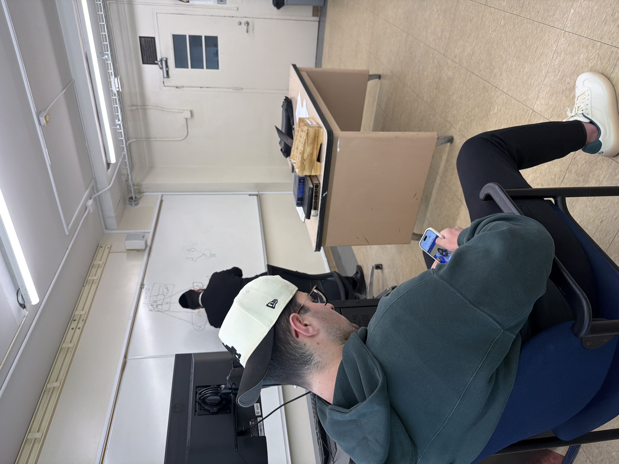 The image shows an interior scene, likely a classroom in the city of New York, United States of America, as inferred from the educational setting. In the foreground, an individual wearing a dark green hoodie, black pants, and white sneakers is seated on a blue chair, engrossed in their mobile phone. The person also sports a white and black baseball cap. In the background, another individual wearing a suit is standing in front of a whiteboard. The environment suggests a classroom setting with a desk and a computer monitor. The room features standard educational design elements such as fluorescent lighting and a neutral color scheme. The overall impression is of an academic or educational environment.