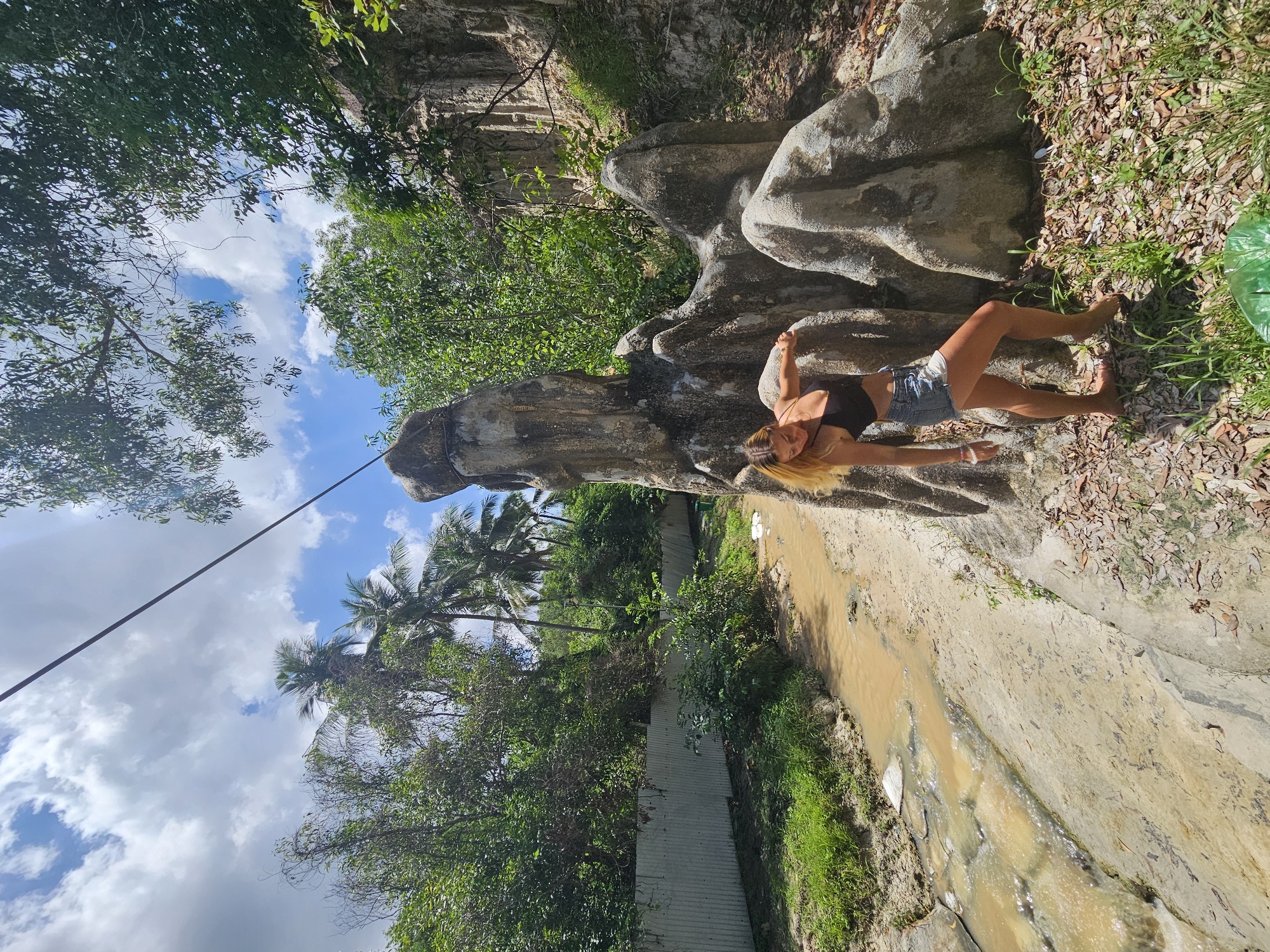 In Mũi Né, Vietnam, a young woman with blonde hair, wearing a black sports bra and denim shorts, poses near a cluster of weathered, eroded rock formations under a bright, cloudy sky. She stands with her left hand on a rock and her body leaning forward. In the background, a muddy stream flows alongside a path, and a corrugated metal fence borders a lush green area with palm trees and other tropical vegetation. The scene suggests a natural landscape altered by human presence, with debris scattered on the ground.