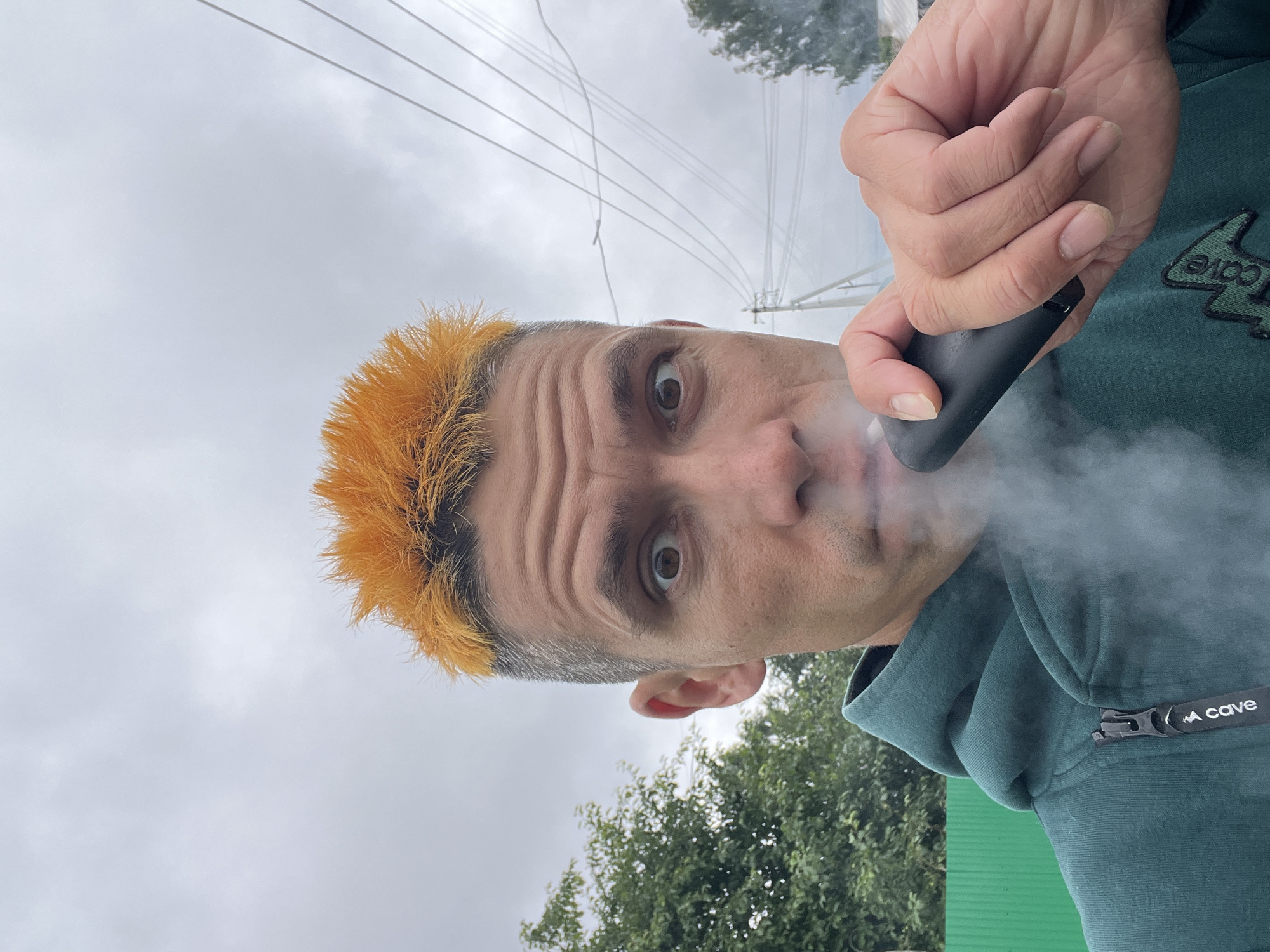 The media file shows a close-up, upside-down selfie of a person with brightly dyed orange hair, exhaling a cloud of vapor. The person has wide, surprised eyes and is wearing a teal hoodie with a black zipper pull that has the text "CAVE" printed on it. They are holding a black vaping device in their right hand. The background is an overcast sky with visible power lines, suggesting an outdoor setting. Green foliage from trees and a green fence are visible at the bottom of the frame. The lighting suggests it could be daytime, but the cloudy sky makes it difficult to determine the exact time. The overall mood conveyed is one of surprise or a captured candid moment.