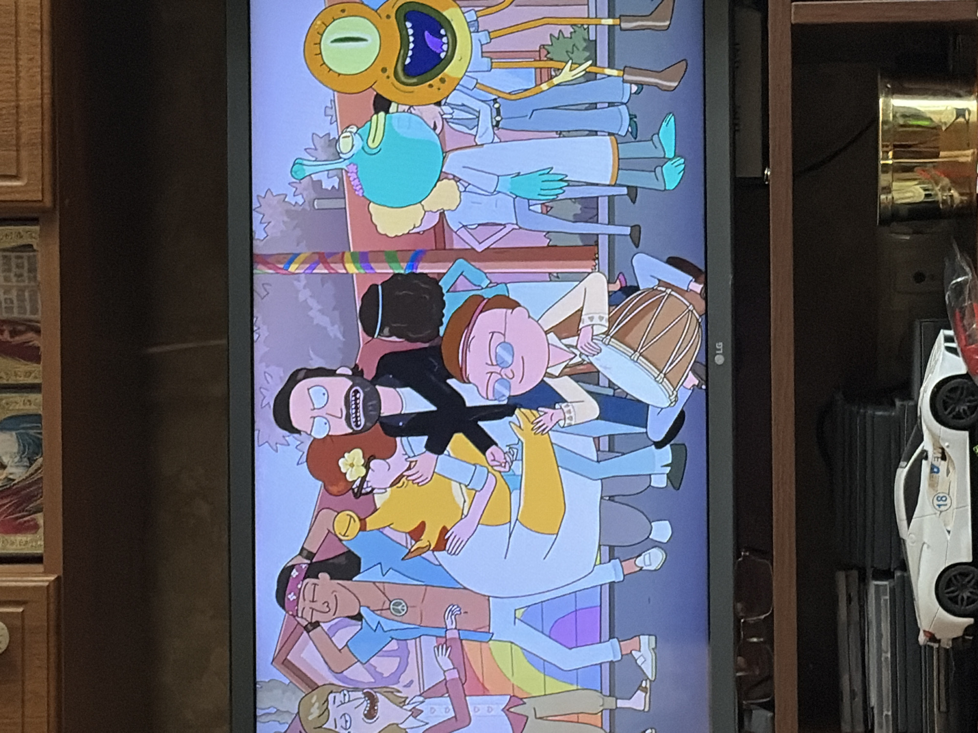 An animated scene displays a group of diverse characters gathered in what appears to be a stylized outdoor environment with a building in the background. The characters are depicted in a cartoonish style, with varying appearances and clothing. One character, a yellow anthropomorphic fox-like creature with large eyes, is being hugged by a human-like character with dark hair. Another human-like character, with an open mouth and wide eyes, stands to the left, while a man with a beard and intense expression is positioned in the center-right. A small, round character with glasses and a hat is seated and playing a drum. Other characters are visible in the background, some with indistinct features. The overall atmosphere seems to be celebratory or social.