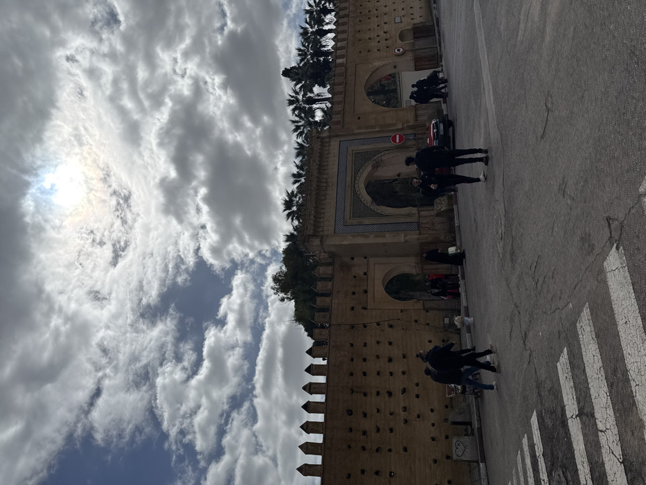 This image depicts a daytime scene in Mechouar Fes Jdid, Morocco, featuring an ancient fortified wall and ornate gateway under a partly cloudy sky. The sky is dominated by large, scattered white and grey cumulus clouds, with the diffused light of the sun visible through a cloud in the upper-center.
The prominent feature is a long, light-brown historical wall stretching across the midground. Its top edge is adorned with crenellated battlements. The wall's surface is marked with numerous small, dark square indentations. A smaller, plain arched entry is located within the wall on the left side. Centrally positioned is a larger, ornate arched gateway featuring intricate geometric tilework in blue, white, and brown hues. To the right of this main gate, a white circular road sign with a red border indicating "No Entry" is visible. Beyond the wall and gates, a line of tall palm trees and other green foliage is visible against the sky.
The foreground consists of a dark grey asphalt road, exhibiting signs of wear. White, cracked pedestrian crossing stripes are visible in the lower-left corner. Approximately 15-20 individuals are present in the scene. On the left side of the road, three men are moving to the right; one is pushing a two-wheeled handcart, and a white bucket is on the adjacent sidewalk. A small white heart outline is spray-painted on the lower part of the wall. In the center-right, two men in dark clothing stand facing each other on the asphalt, appearing to be engaged in conversation. A dark-colored sedan is partially visible behind them. Further right, a group of 5-7 individuals is congregated near another opening in the wall. Individuals are generally observed walking, standing, or interacting in small groups.