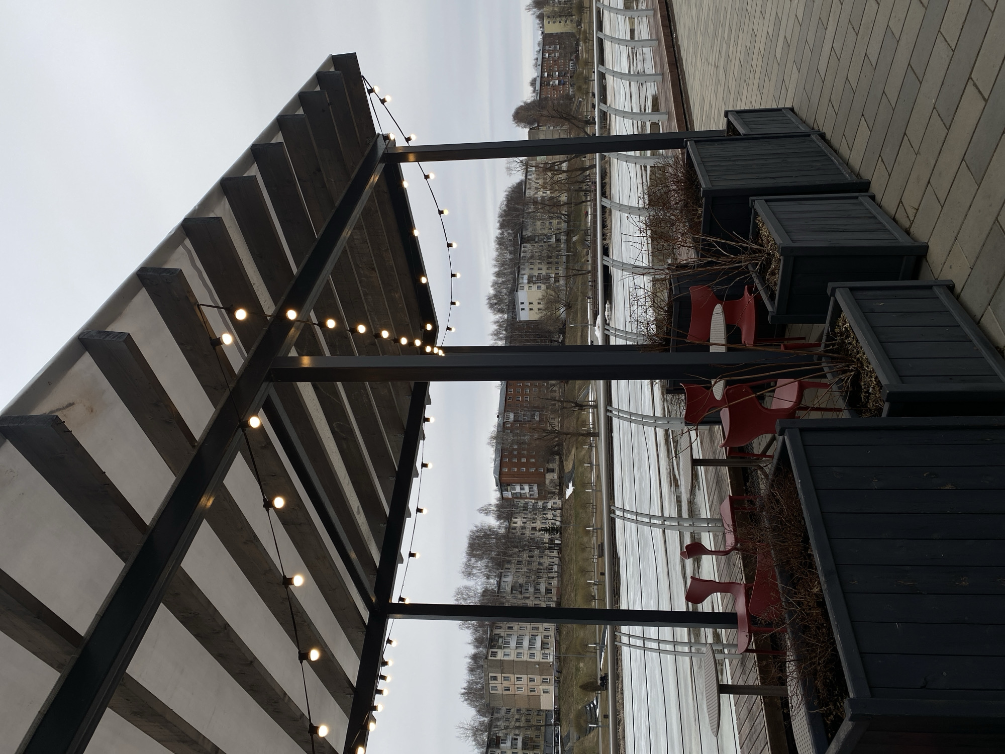 The image depicts an outdoor dining area, likely a cafe or restaurant, in Votkinsk, Russia. The location overlooks what seems to be a waterway or riverbank. A metal-framed canopy structure with strung lights covers part of the dining area. Several red chairs and tables are visible, positioned on a wooden deck. Square black planters line the edge of the deck, containing dry vegetation. Across the waterway, multi-story apartment buildings characteristic of Russian urban landscapes are visible under an overcast sky. The ground appears to have patches of snow or ice, suggesting a cold or late-winter setting. The walkway next to the planters is paved with interlocking stones. A curved metal railing borders the deck near the waterway. No people are visible in the frame.