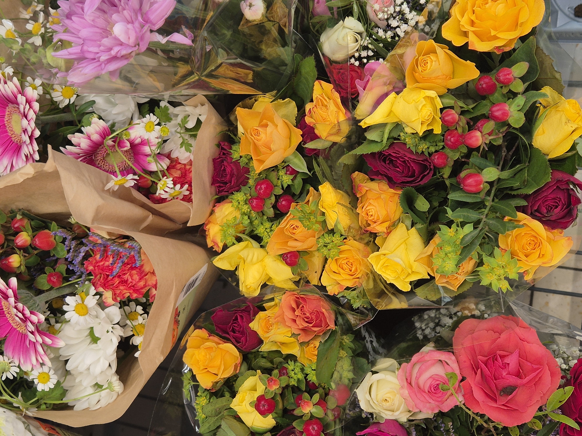 An overhead photograph displays a dense arrangement of multiple flower bouquets, likely a retail display, in Unknown, Unknown. The scene is dominated by a variety of cut flowers including roses in yellow, orange, deep red, light pink, and white hues. Prominent among them are pink and white gerbera daisies, pinkish-purple and white chrysanthemums, and small white daisies with yellow centers. Accompanying flora includes clusters of small, bright red berries (resembling hypericum), green foliage, and sparse purple filler flowers. Several bouquets are encased in natural brown kraft paper, while others are individually wrapped in clear plastic sleeves that exhibit reflections. The bouquets are tightly grouped on a dark surface, possibly a display shelf or cart, with faint grid patterns visible through some of the transparent packaging. No people or actions are depicted.