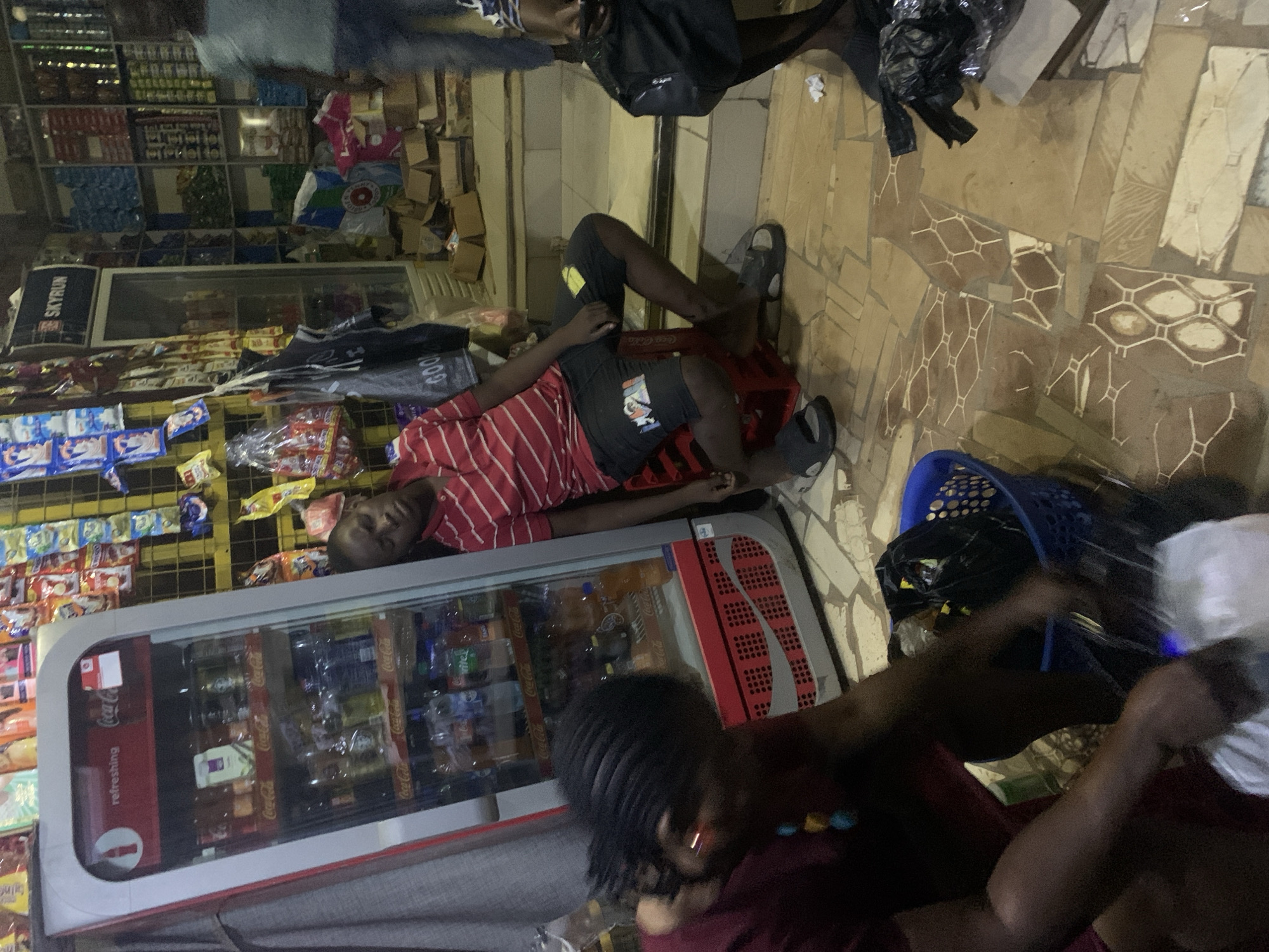In an indoor shop, a young Black male in a red and white striped shirt and black shorts sleeps leaning against a red plastic crate. Beside him, a blue plastic basket contains black plastic bags. A Black female with dark braided hair is kneeling in the foreground, handling white plastic bags. Behind them, a refrigerator is filled with various drinks. Shelves stocked with goods line the walls, and cardboard boxes are stacked near the entrance. The floor is tiled with a patterned design.