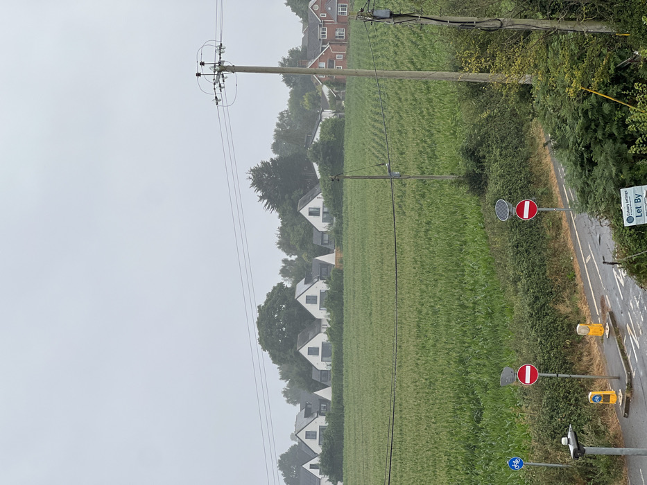 The image displays an overcast rural-urban fringe landscape in the Tithebarn area of Exeter, United Kingdom. In the foreground, a wet asphalt road or path runs horizontally, bordered by green foliage. On the left side of the path, a paved section features a blue circular sign with a white bicycle symbol, two yellow bollards, and a street light pole. A red circular 'No Entry' sign is centrally positioned on this paved area. Further right, another red circular 'No Entry' sign with a solar panel mounted above it stands between the path and the field. A "Let By" sign for "Estuary Lettings" is partially visible in the bottom right corner.
The midground is dominated by a large, cultivated field of dense green crops, likely corn or maize, arranged in visible rows receding towards the background. Overhead power lines traverse the upper portion of the image, supported by two wooden utility poles; a thicker pole with electrical equipment is on the right, and a thinner pole is further left within the field. In the background, a line of mature trees partially obscures and frames a residential area. Several modern, white-rendered houses with dark, steep gabled roofs and prominent dark-framed windows are visible behind the field. To the far right, a traditional red-brick house with a dark roof is also present. The sky is uniformly light grey and overcast. No individuals, animals, or vehicles are present in the scene.