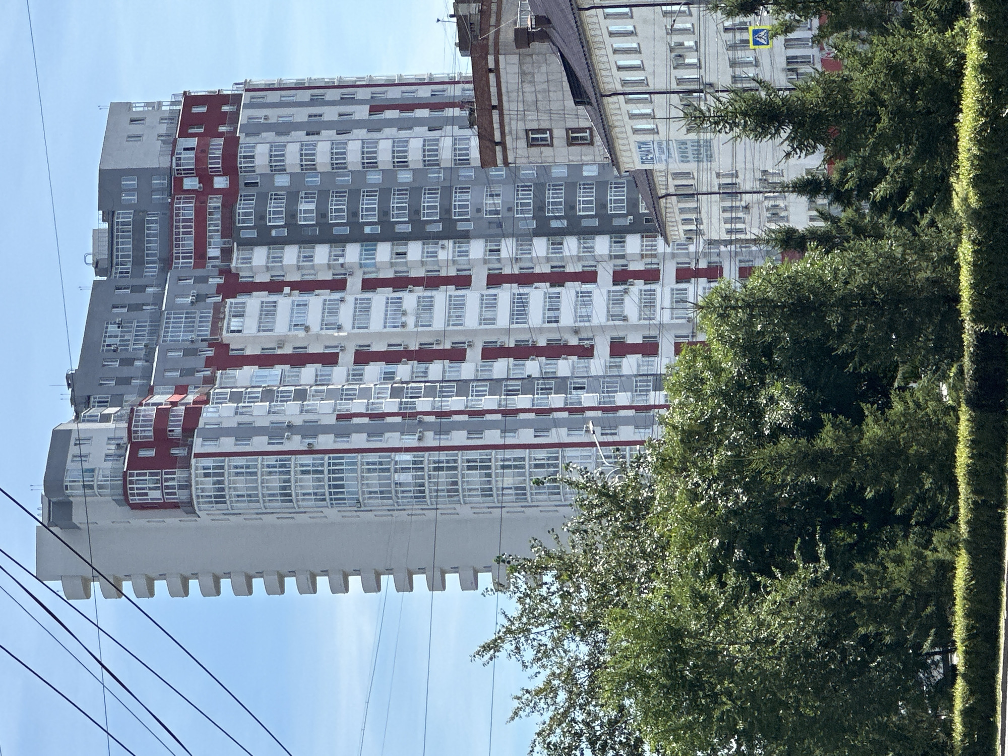 A tall, modern residential building with a white, grey, and red facade dominates the center of the frame. The building features numerous balconies and large windows. In the foreground, a line of green trees and bushes obscures the lower portion of the building and adjacent structures. A clear blue sky is visible above. Several dark power lines traverse the upper portion of the image. To the right of the main building, another, older, light-colored building with a dark roof is partially visible. A street sign with a pedestrian symbol is visible near the older building. The scene appears to be an urban environment during daylight.