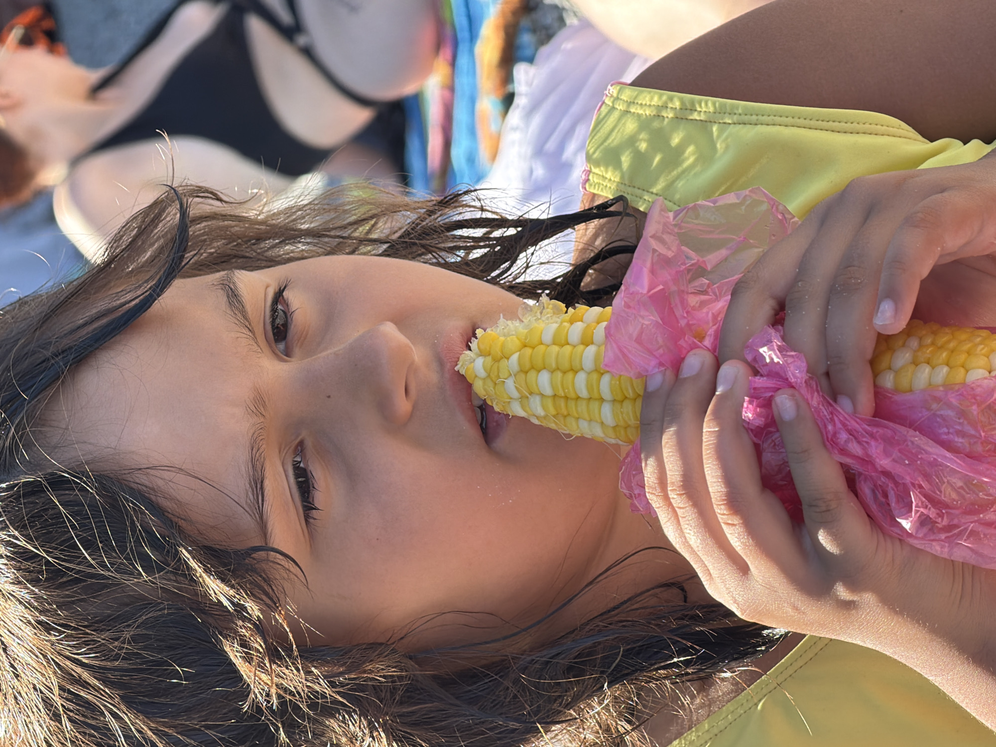 In this close-up, low-angle shot, a young person with dark, wet hair is lying down, holding a cob of corn wrapped in pink plastic. The person is wearing a yellow bikini top. Their face is turned towards the camera, with one eye visible and a slight smile on their lips. The corn is partially eaten, with kernels visible on the cob. In the background, another person is visible, lying down and wearing a black bikini. The scene appears to be outdoors, possibly at a beach or park, given the lighting and casual attire. The sunlight suggests it is daytime, and the relaxed posture of the subjects implies a leisurely activity. The backdrop includes colorful fabrics, adding to the casual, summery atmosphere. No text is visible in the image. The context provided places the scene in Partenit, Ukraine.