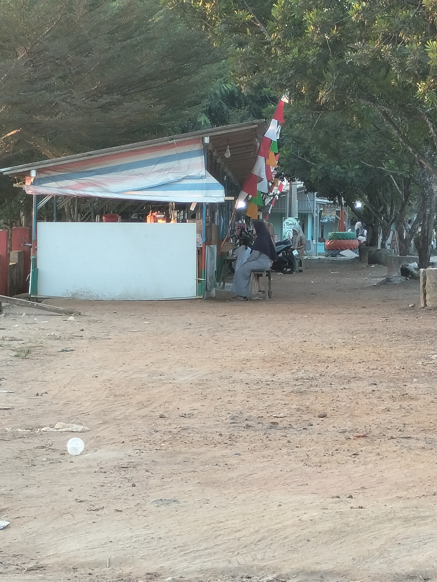 In the city of Karang Indah, Indonesia, a scene unfolds on a dirt path near a roadside stall. A woman wearing a hijab and a long skirt sits on a wooden stool, her gaze directed towards the left. Beside her, a motorcycle is parked. The stall itself features a striped awning and a large white board, likely for display or advertising. Further in the background, other structures and trees are visible, suggesting a semi-urban or rural setting. The ground is covered in sand and small pebbles, with a white plastic cup lying on the ground in the foreground. The overall impression is one of a humble marketplace or rest stop.