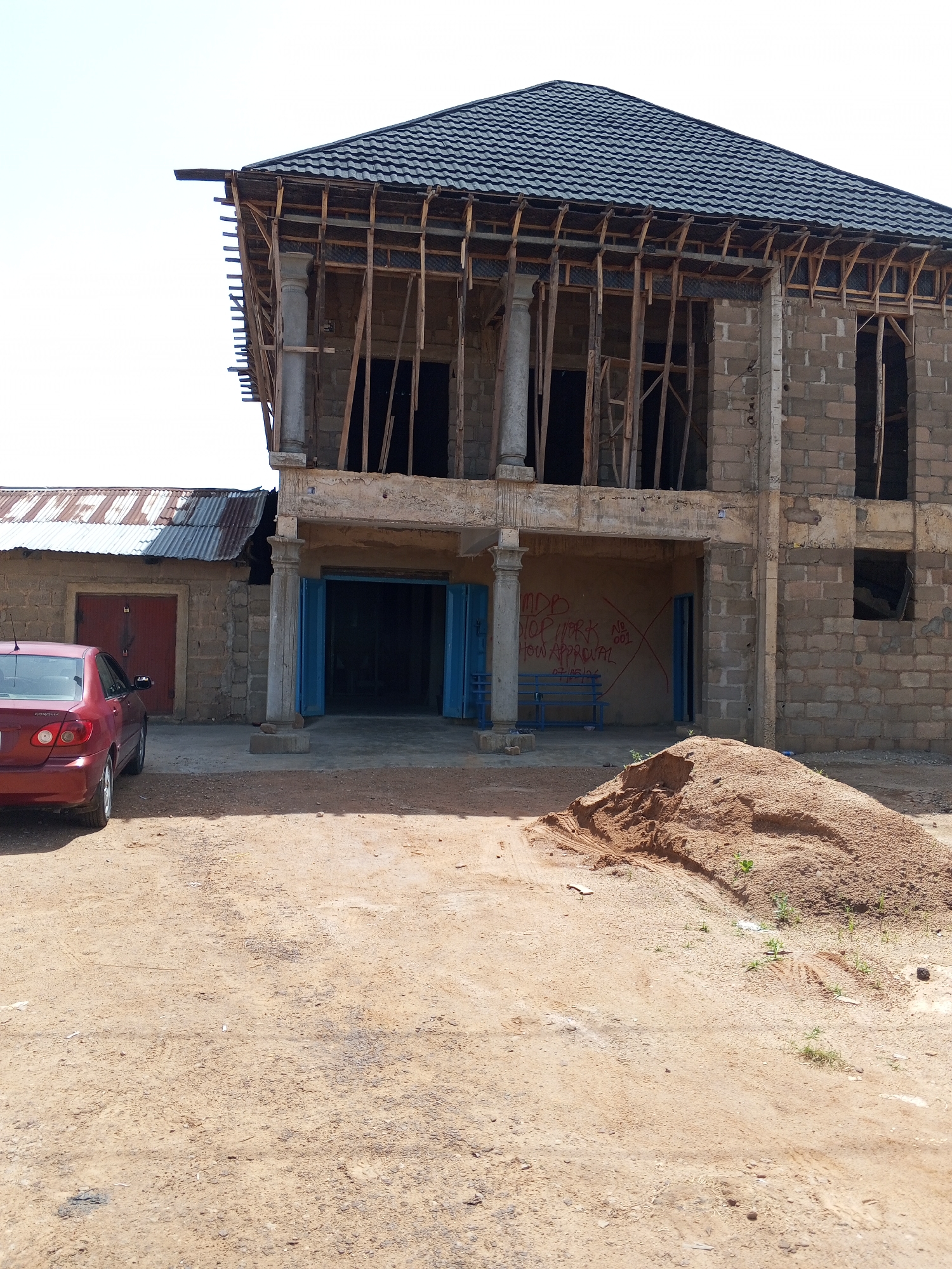 A construction site in Bukuru, Nigeria features a two-story building under construction. The building has a dark, tiled roof and exposed wooden framing for the upper floors. The ground floor has large, open doorways with blue doors, and a covered porch area supported by pillars. Red graffiti on the wall of the porch reads "MDB STOP WORK NO 001 HOW APPROVAL 07/05/2024".
To the left of the main building is a smaller, single-story structure with a corrugated metal roof. A red sedan is parked in front of this structure. A large pile of sand is situated in the foreground, indicating ongoing construction work. The ground is dirt and appears dry. The sky is clear and bright, suggesting it is daytime. The scene appears to be an active construction zone in a rural or semi-rural Nigerian setting.