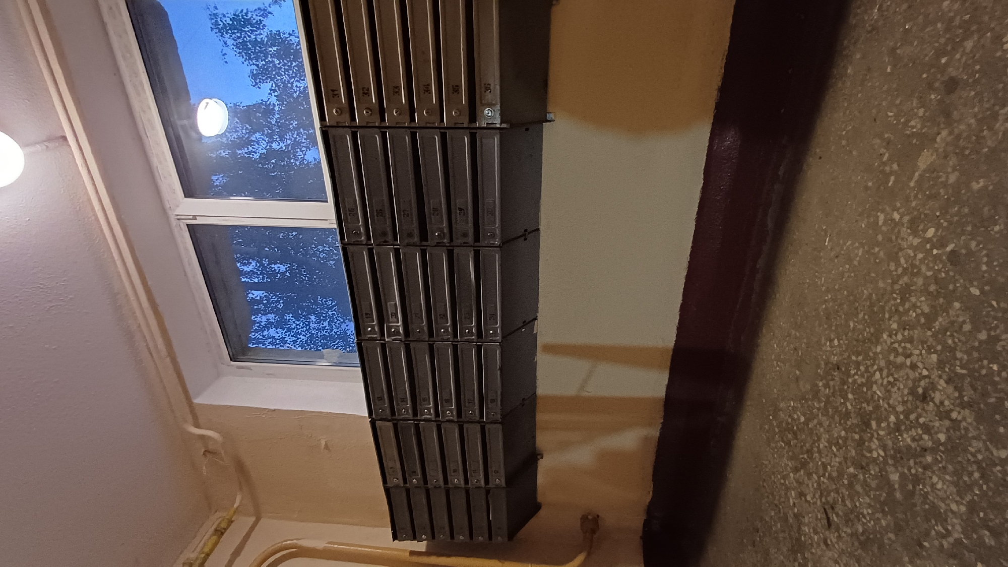 The image shows an indoor hallway in Balashikha, Russia. A stack of metal mailboxes, arranged in four rows, is mounted on the wall. To the left of the mailboxes is a window showing a view of the sky and tree branches. Beneath the window, pipes run along the wall. The wall to the right of the mailboxes is beige with a dark red stripe. The floor is speckled beige.