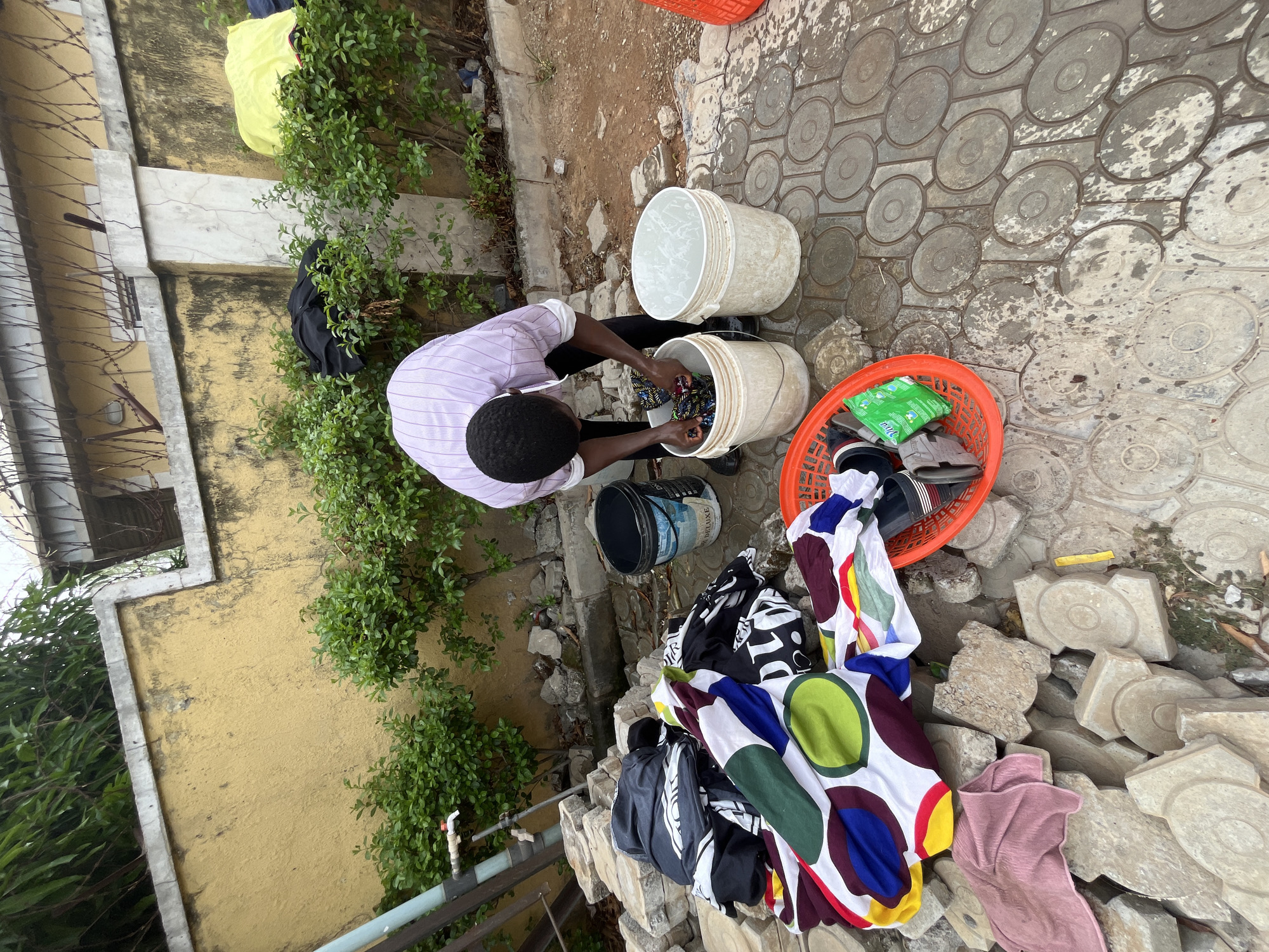 A man is washing clothes outdoors. He is wearing a purple and white striped shirt and black pants. He is standing over two white buckets, one filled with clothes and the other with water. Next to him is an orange basket with shoes and a green detergent package inside. A black bucket and a pile of clothes are nearby. The ground is paved with patterned concrete tiles. The location is likely within the city of Abuja, Nigeria. A building with a yellow wall and a green bush is visible in the background.