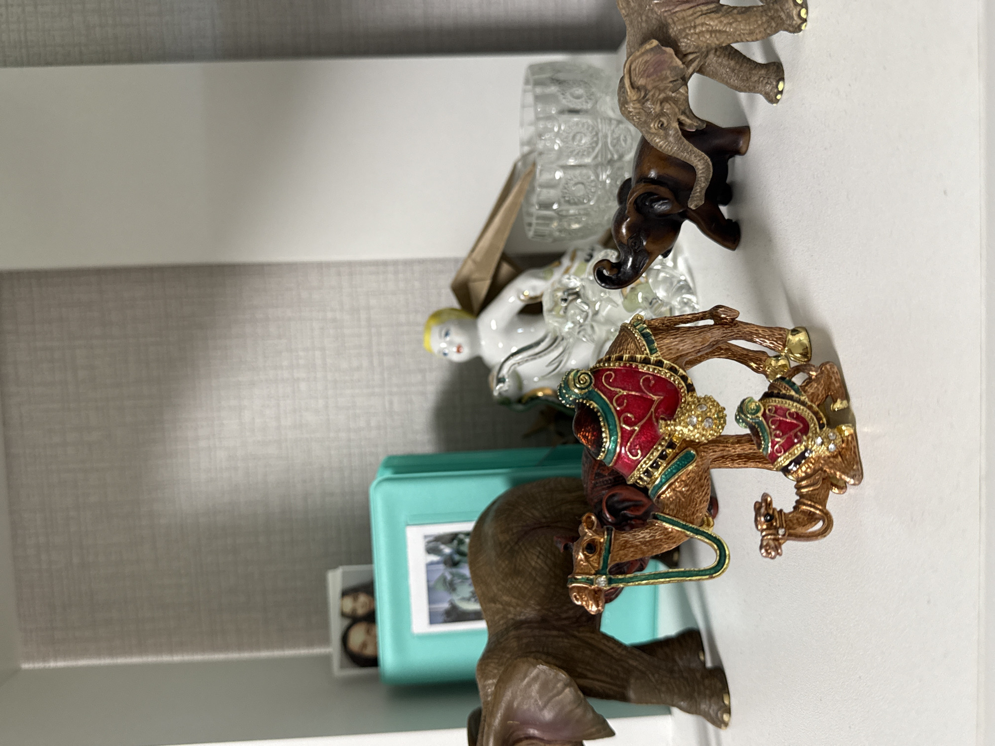 The image displays a collection of decorative figurines arranged on a shelf. The subjects include several elephant figurines in varying sizes and colors, with one larger elephant in the foreground. There is also a decorative horse figurine adorned with red and gold ornamentation, and a figurine of a person. Interspersed among the figurines are a clear glass decorative item and a teal-colored box. A small framed photograph is visible behind the teal box, showing two faces. The scene is set on a shelf with a textured gray background wall. The lighting suggests an indoor setting, likely a room in a home or office. There is no indication of the time of day, weather, or any specific emotions conveyed by the arrangement. No visible text is present in the image.