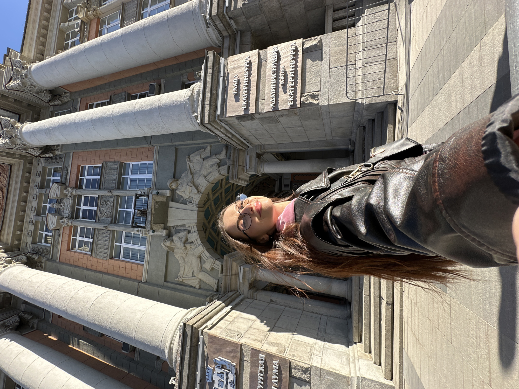 A daytime selfie shows a woman in a black leather jacket and eyeglasses standing in front of a large, ornate building. Her long brown hair is visible. She has a pink scarf or shirt underneath the jacket. The building behind her has large columns and detailed architectural elements. Inscriptions in Russian are on the building, including "Дума Екатеринбурга" (Yekaterinburg Duma) and "Администрация города Екатеринбурга" (Administration of the City of Yekaterinburg), indicating the location. The facade includes sculptural figures and rectangular windows. The building appears to be made of stone or concrete.
