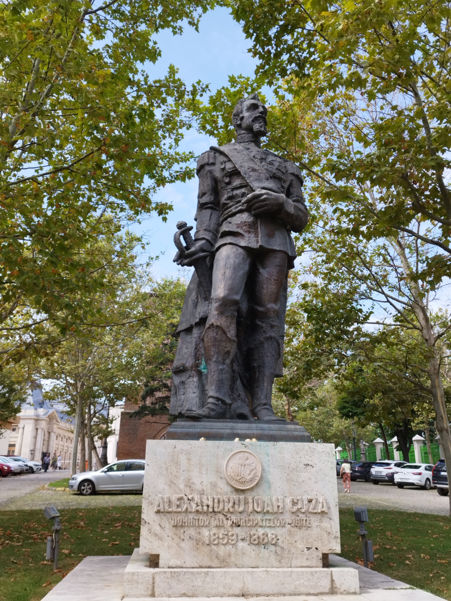 This image shows a bronze statue of Alexandru Ioan Cuza, the first ruling prince of Romania, situated in a park-like setting in Bucharest, Romania. The statue stands on a marble pedestal inscribed with "ALEXANDRU IOAN CUZA DOMNITOR AL PRINCIPATELOR UNITE 1859-1866". The statue depicts Cuza in military attire, holding a sword in his left hand and a scroll in his right.
The background features lush green trees with some autumn foliage, a clear blue sky, and several parked cars. To the left of the statue, a grand building with classical architecture is visible, suggesting it might be a significant historical or governmental institution. In the distance, a green metal fence encloses another area. A few people are seen walking in the background, some near the cars and others near the building. The overall scene appears to be a public space, likely a monument or a historical site within the city.