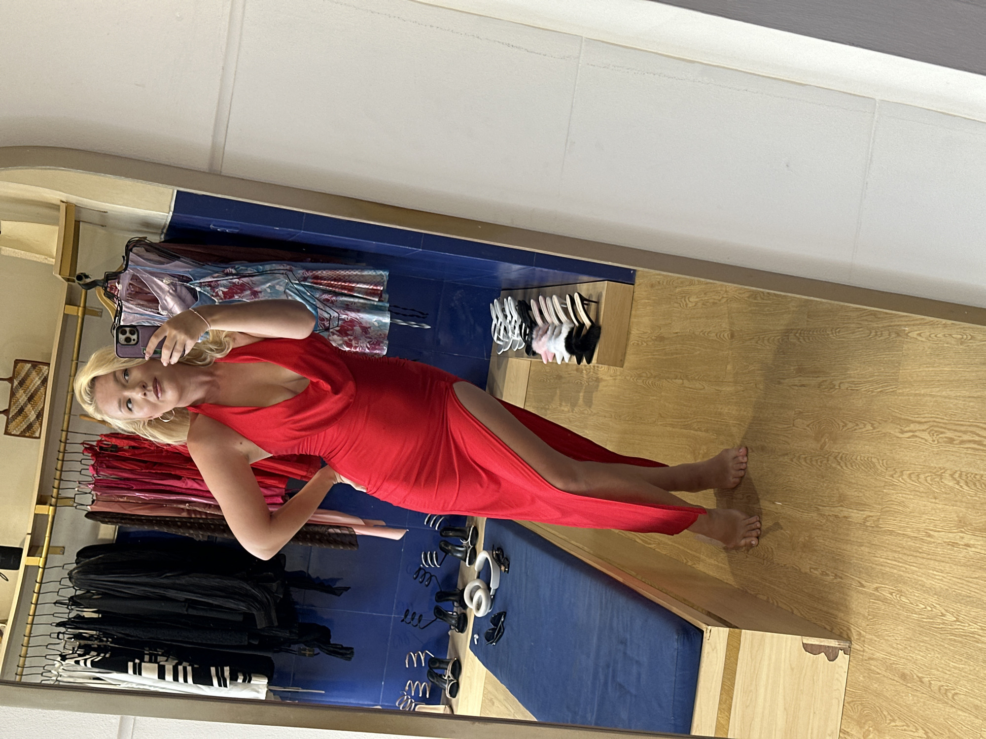 A woman stands in a dressing room, reflected in a mirror. She holds a smartphone in her left hand, presumably taking a photo. She is wearing a red, floor-length dress with a high slit. Her blonde hair is pulled back, and she is wearing hoop earrings.
Behind the woman is a clothing rack with various garments, ranging from black dresses to pink and red tops. Below the clothing rack are shoes and accessories on display. The floor is wood-patterned. The walls are white and blue.
This scene appears to be a dressing room in a retail clothing store, likely located in North Kuta, Indonesia.