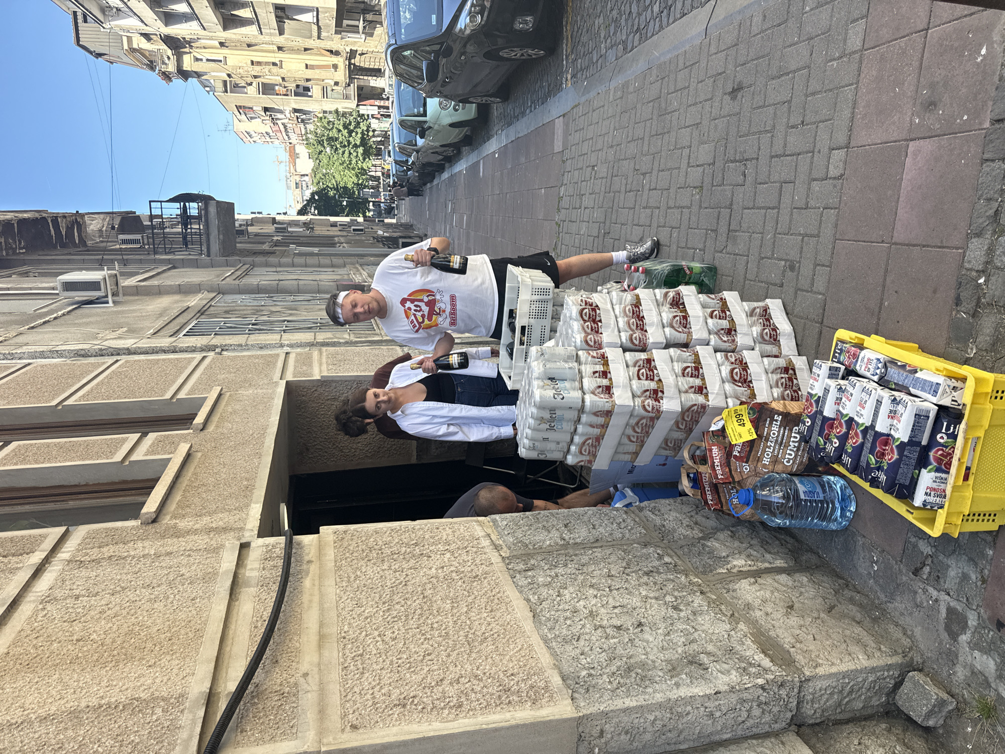 The scene is set on a sidewalk in Belgrade, Serbia, featuring a man and a woman standing near a stack of goods. The man, wearing a white t-shirt with a "BetBoom" logo, black shorts, a white headband, and grey sneakers, holds two champagne bottles. Next to him is a stack of what appear to be beer or beverage cases. The woman, wearing a white shirt, is seen standing behind him. Another person appears to be sitting by the stack of goods.
To the right of the stack, there is a yellow plastic crate containing juice boxes and other items. A bottle of water and a bag of wood coal are placed in front of the crate. The sidewalk is made of interlocking grey brick.
In the background, several cars are parked along the side of the street, including a dark grey car and a green Fiat 500. Buildings are visible on both sides of the street.