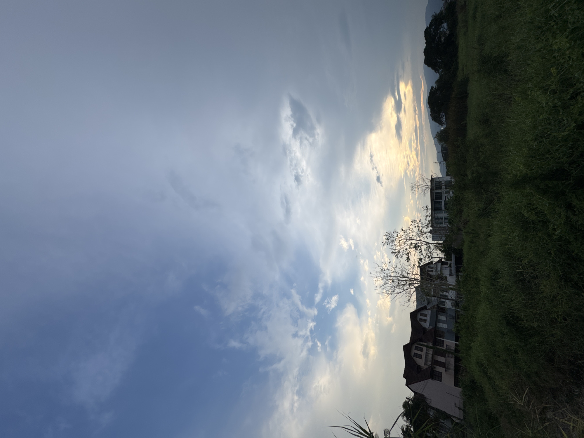 The image shows a view in บ้านกาญจน์กนก 23, Thailand. The sky is cloudy, with patches of blue sky visible. Buildings, including residential houses, are visible amidst tall grass and foliage. In the background, there's a suggestion of distant hills or mountains along the horizon. A bare tree stands near the buildings.