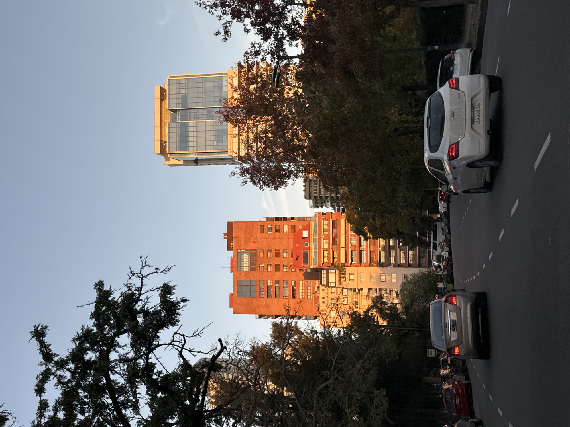 An upward-tilted perspective captures a street scene in Buenos Aires, Argentina, featuring two prominent buildings illuminated by the setting sun. The building on the left is a tall, modern structure with a gold-colored exterior and reflective glass windows. To the right of it, a shorter building with an orange facade displays rows of windows, some reflecting the sunlight.
The foreground is filled with trees, their branches extending towards the buildings. A row of parked cars lines the side of the road, with a white car prominently visible on the right. The asphalt road is divided by painted lines, indicating lanes. The sky above is a clear blue. The overall scene is peaceful, bathed in the warm glow of the evening light.
