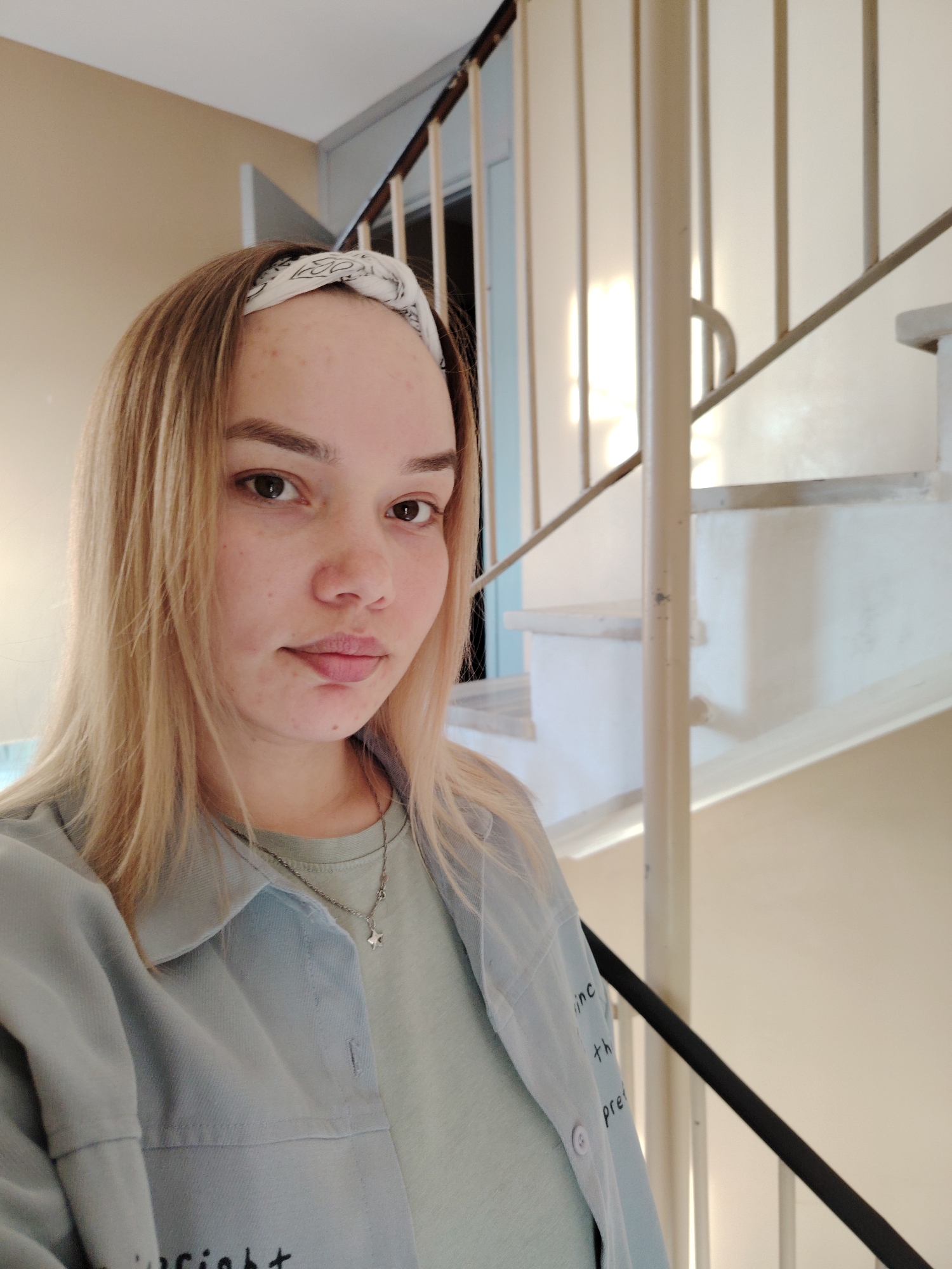 A young woman with blonde hair and a white bandana stands in front of a spiral staircase. She is wearing a light green t-shirt and a light blue button-down shirt with text printed on the sleeve. The staircase is painted white with a black metal railing. The walls are beige. The location is Марковский, Russia.