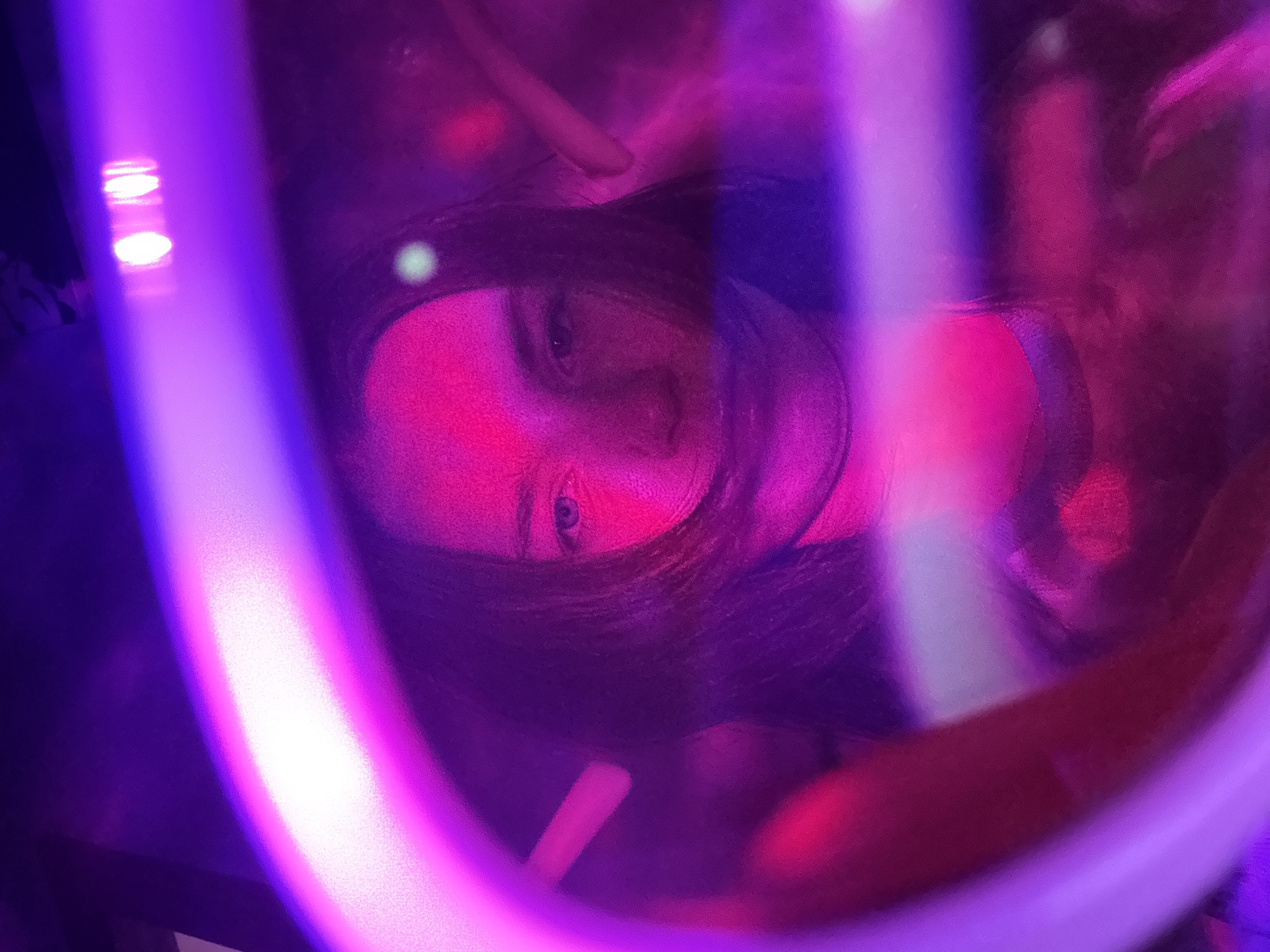The image is a vibrant, close-up portrait featuring a person, likely female, framed and partially obscured by prominent, blurry, glowing bands of purple and pink-red light in the foreground. Her face is illuminated by strong magenta and purple light, with her right eye clearly visible, looking directly towards the viewer. Dark hair frames her face.
The scene depicts an indoor setting, likely a nightclub, bar, or entertainment venue, given the intense artificial lighting. The time of day is night. The glowing foreground elements appear to be neon tubes or LED strips, creating a tunnel-like or abstract framing effect around the subject. In the background, softer, blurred lights in white, red, and purple hues are visible, contributing to the atmospheric, moody ambiance. The person appears to be posing for a photograph or selfie within this colorful environment. There is no visible text in the image.