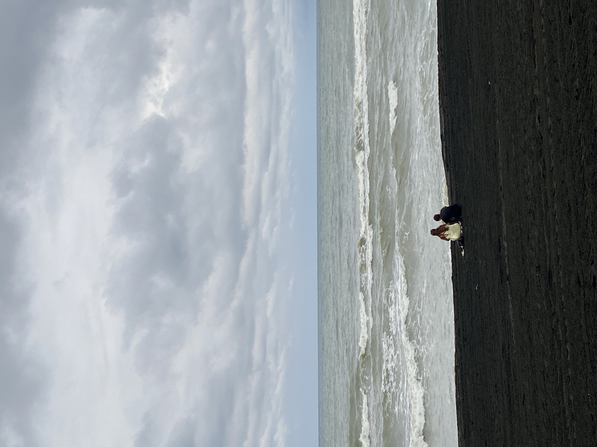 A couple sits on a black sand beach facing the sea under a cloudy sky. The woman wears a light-colored dress and the man wears a dark shirt. Waves crash on the shore. The scene is located on the coast of Ureki, Georgia.