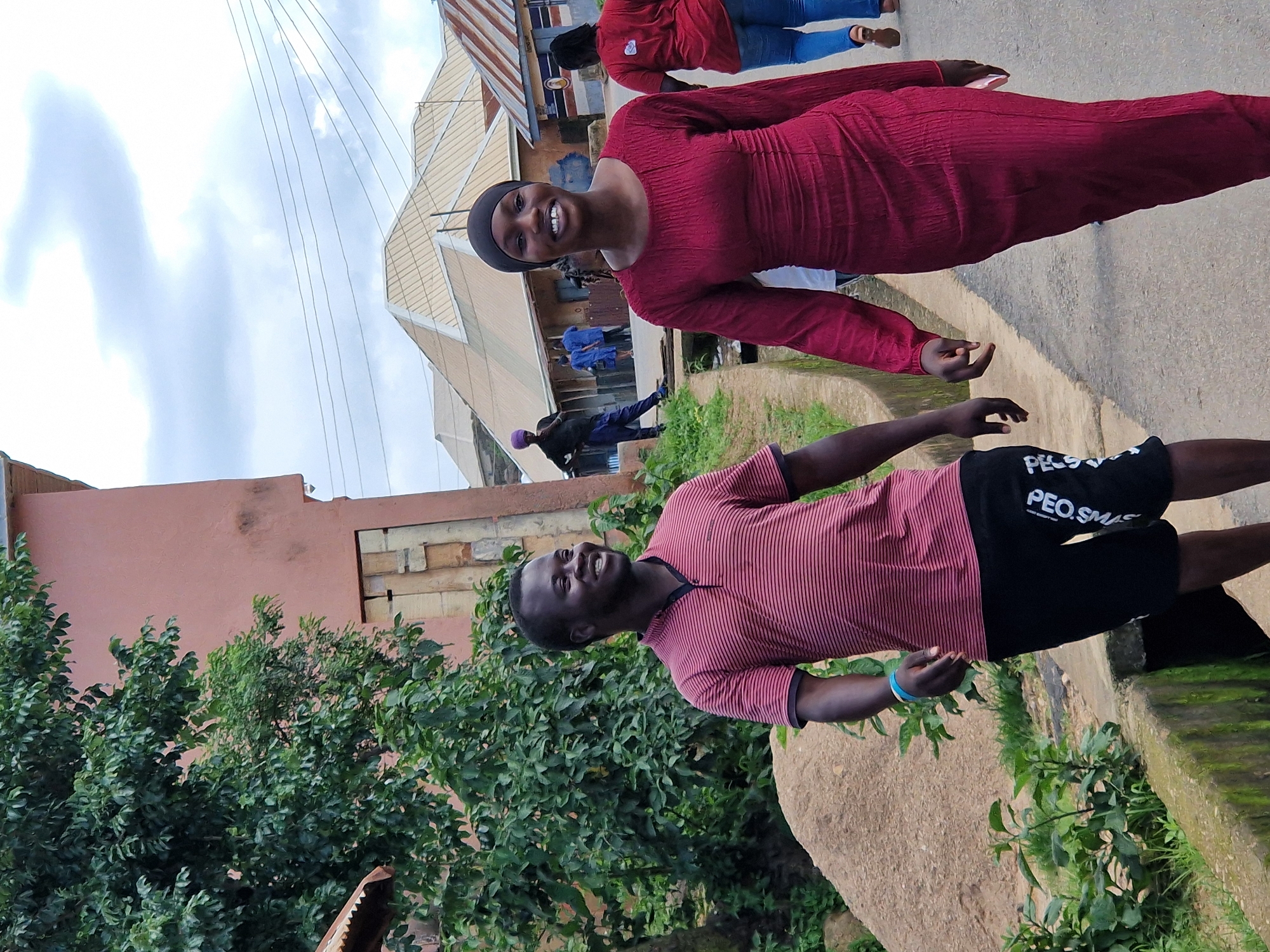 A young Black man and a young Black woman walk side-by-side on a paved street. The man wears a red and pink striped polo shirt and black shorts with white lettering. He has a blue bracelet on his left wrist. The woman wears a long-sleeved, dark red, textured dress and a black headscarf. They are both smiling and looking forward. Behind them, a red-shirted woman in blue jeans walks away from the camera. In the background, several buildings with corrugated metal roofs are visible, as are electric wires strung across the sky. Trees and vegetation line the left side of the street, and a concrete drain runs along the edge of the road. The sky is overcast with clouds.
