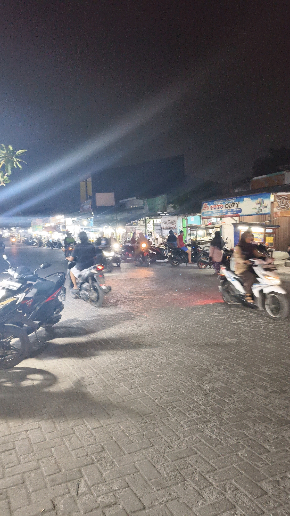 A nighttime scene in Satriajaya, Indonesia, depicts a street bustling with motorcycle traffic. The road surface is paved with grey interlocking bricks. In the background, a row of small shops and stalls are illuminated, with signs indicating businesses such as "FOTO COPY". Several individuals are visible riding motorcycles, with some passengers also present. The lighting is a mix of artificial light from the shops and headlights from the vehicles, creating illuminated streaks and shadows. The sky is dark, suggesting it is nighttime. A palm tree is partially visible on the left side of the image.