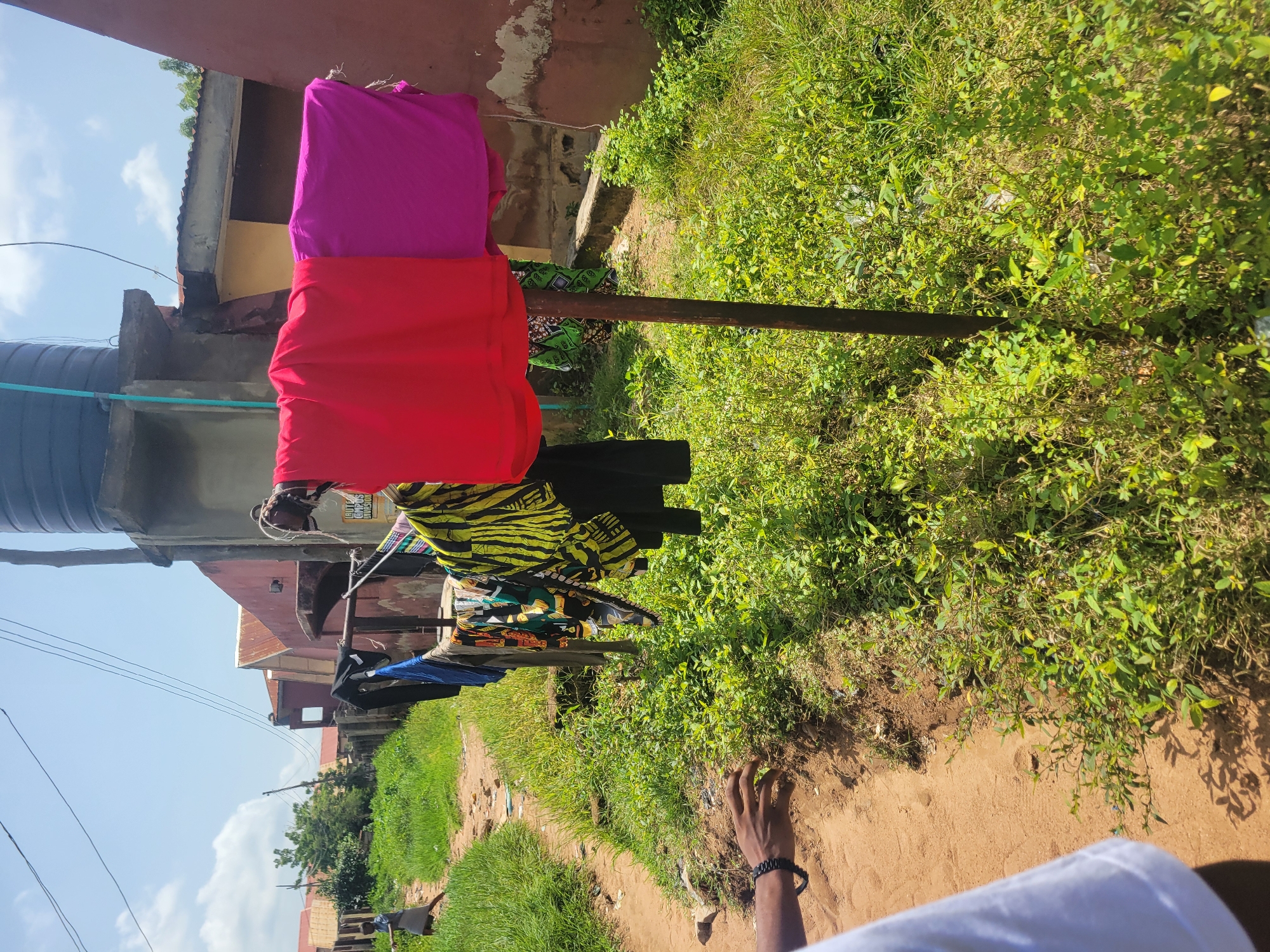 In Ogbomosho, Nigeria, on a sunny day, laundry hangs on a clothesline supported by a wooden pole. Two colorful pieces of fabric, one red and one magenta, are prominently displayed on the line. Various other garments, including a black garment, a patterned green and black garment, and a blue garment, are also present. To the left, a person's arm and hand, adorned with a black bracelet, are visible. A dirt path runs alongside a patch of green grass and bushes. In the background, buildings with red roofs and a water tank are visible under a clear blue sky with scattered clouds. A person wearing a grey dress is walking on the dirt path in the distance.