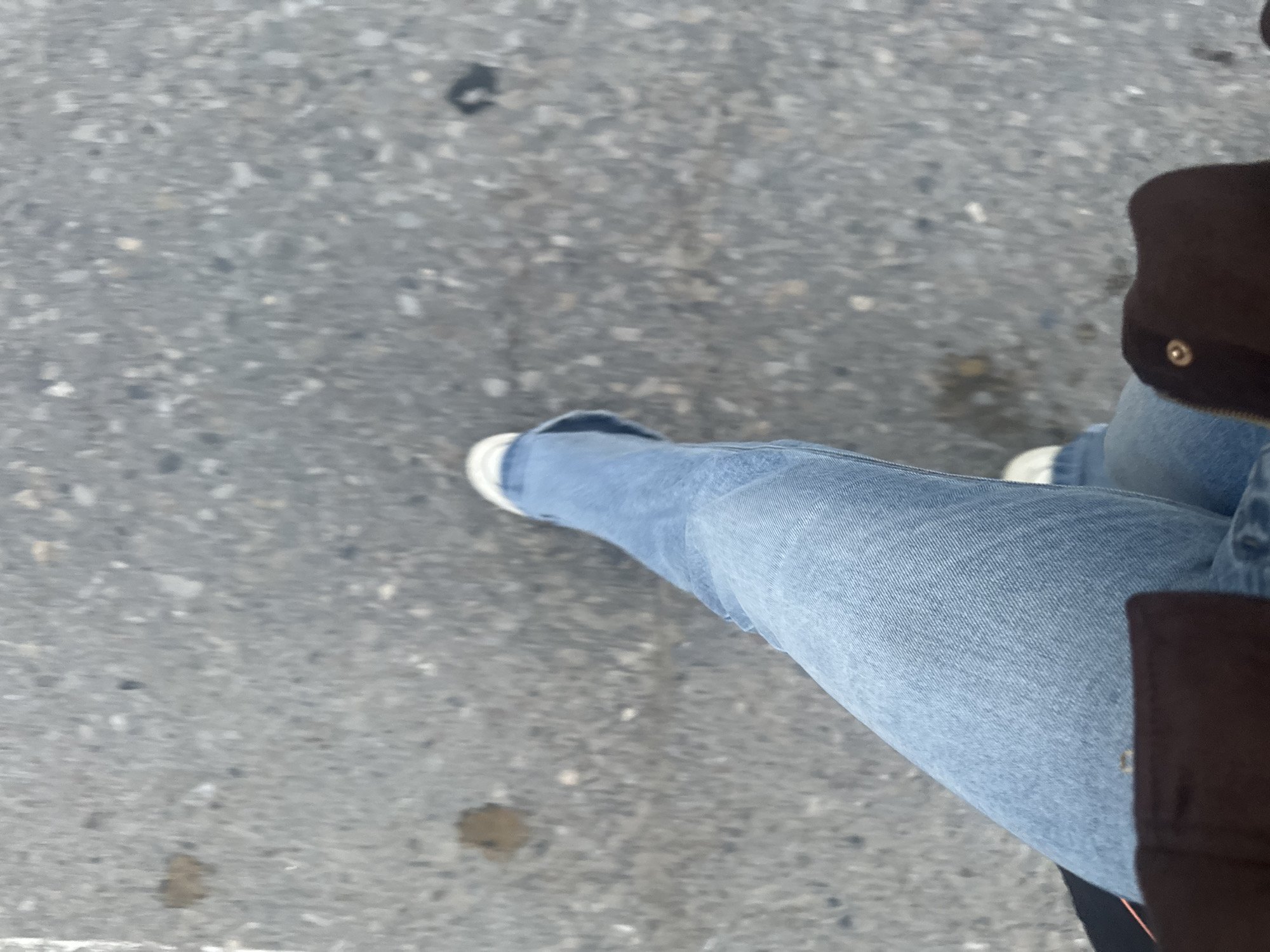 An overhead shot depicts a person's legs on a grey, speckled asphalt surface. The person wears light-wash blue jeans and white sneakers. A brown jacket is partially visible on the right side of the frame. The setting appears to be outdoors, likely a street or sidewalk in Высокий, Russia, given the location context.