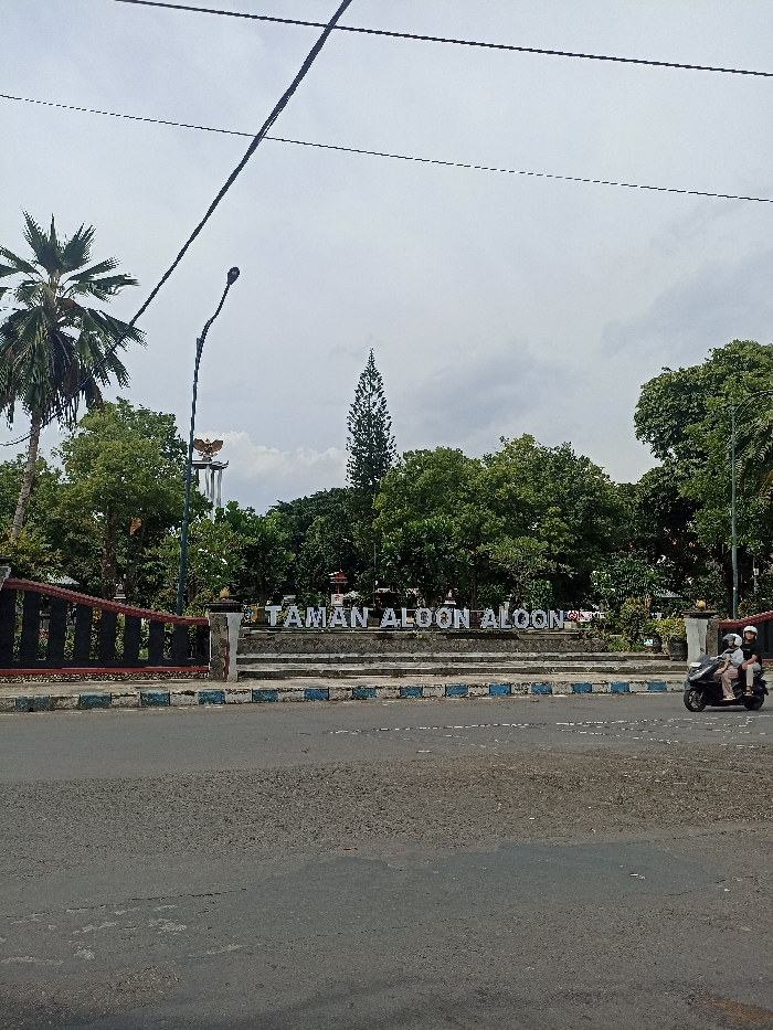 The image displays an outdoor scene of "Taman Aloon Aloon" (Alun-alun Park) in Tulungagung, Indonesia, under an overcast sky. In the foreground, a paved road is bordered by distinctive blue and white striped curb stones.
A prominent sign in white block letters, "TAMAN ALOON ALOON," marks the entrance to the park, situated on a raised stone platform with steps. Behind this sign, the park is characterized by lush green trees and foliage. A tall palm tree stands on the far left, and a slender, conical evergreen tree is visible towards the center-right.
In the mid-ground, a monument or statue, appearing to be a golden eagle-like figure (likely the Garuda, Indonesia's national emblem), is partially visible atop a structure amidst the trees. A tall, dark street light curves over the park entrance. Above, power lines stretch across the light grey, cloudy sky.
On the road to the right, two individuals are riding a black scooter. Both are wearing helmets, indicating a typical daytime activity. The overall setting portrays a public park area within a town, with no specific event taking place, but rather everyday life.