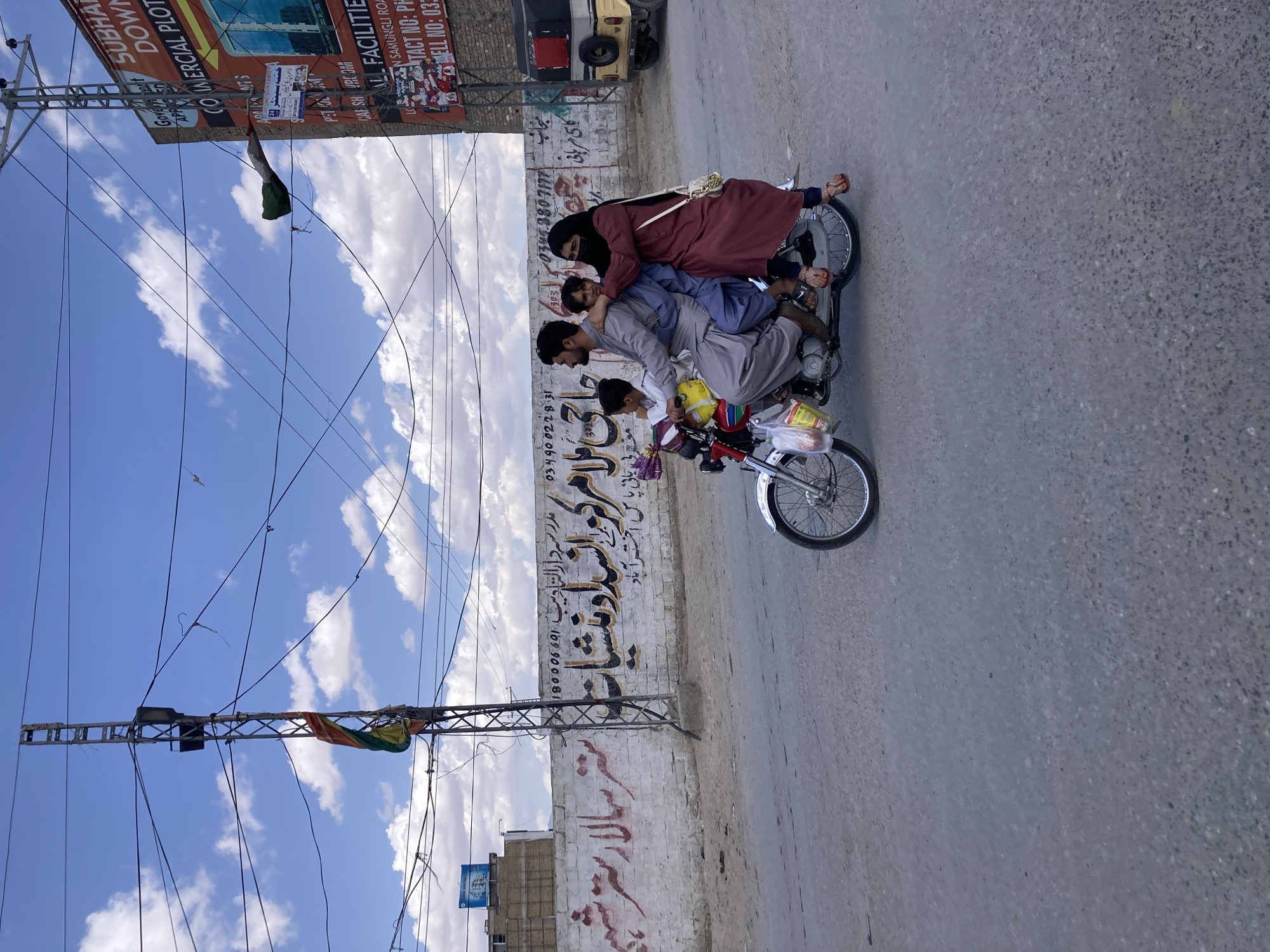 The image depicts a family of four riding on a single motorcycle on a street in Quetta City Tehsil, Pakistan. A man is driving the motorcycle, with a child in front of him. Behind the man, another man is seated, and a woman wearing a hijab is seated behind him. The motorcycle is laden with a bag tied to the front and various other items. The background consists of buildings with Urdu script on the walls, utility poles with power lines, and commercial billboards. The street is paved and appears to be an average urban road. The sky above is blue with scattered clouds.