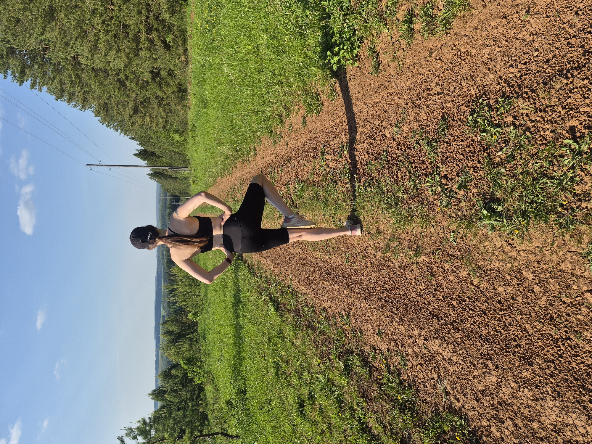 A woman in black athletic attire stands on a dirt path, hands on hips, in an area with green grass and trees. Power lines are visible in the background. The sky is blue with some clouds.