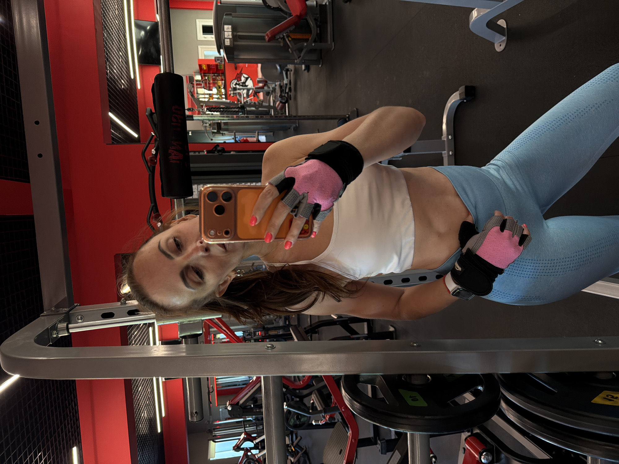 A woman is captured in a mirror selfie within an indoor gym in Birobidzhan, Russia. She is the central subject, holding a smartphone with an orange case to photograph her reflection. She wears a white sports bra or crop top, light blue patterned leggings, and pink and black fingerless workout gloves. A smartwatch with a white strap is visible on her left wrist.
The gym environment features prominent red walls, a dark floor, and bright linear lighting fixtures. Various pieces of gym equipment are visible, including a large silver/grey machine frame she stands near. In the background, reflections and direct views show other gym apparatus such as benches, weight racks, and stacked weights. Black weight plates bear the numbers "10" (on a green label) and "40" (on a yellow label). A padded handle on one of the machines has partially visible text, appearing to read "DEM MAX". Her expression is neutral to slightly smiling, suggesting she is comfortable or pleased during her workout break. The time of day is not indicated by the indoor setting.