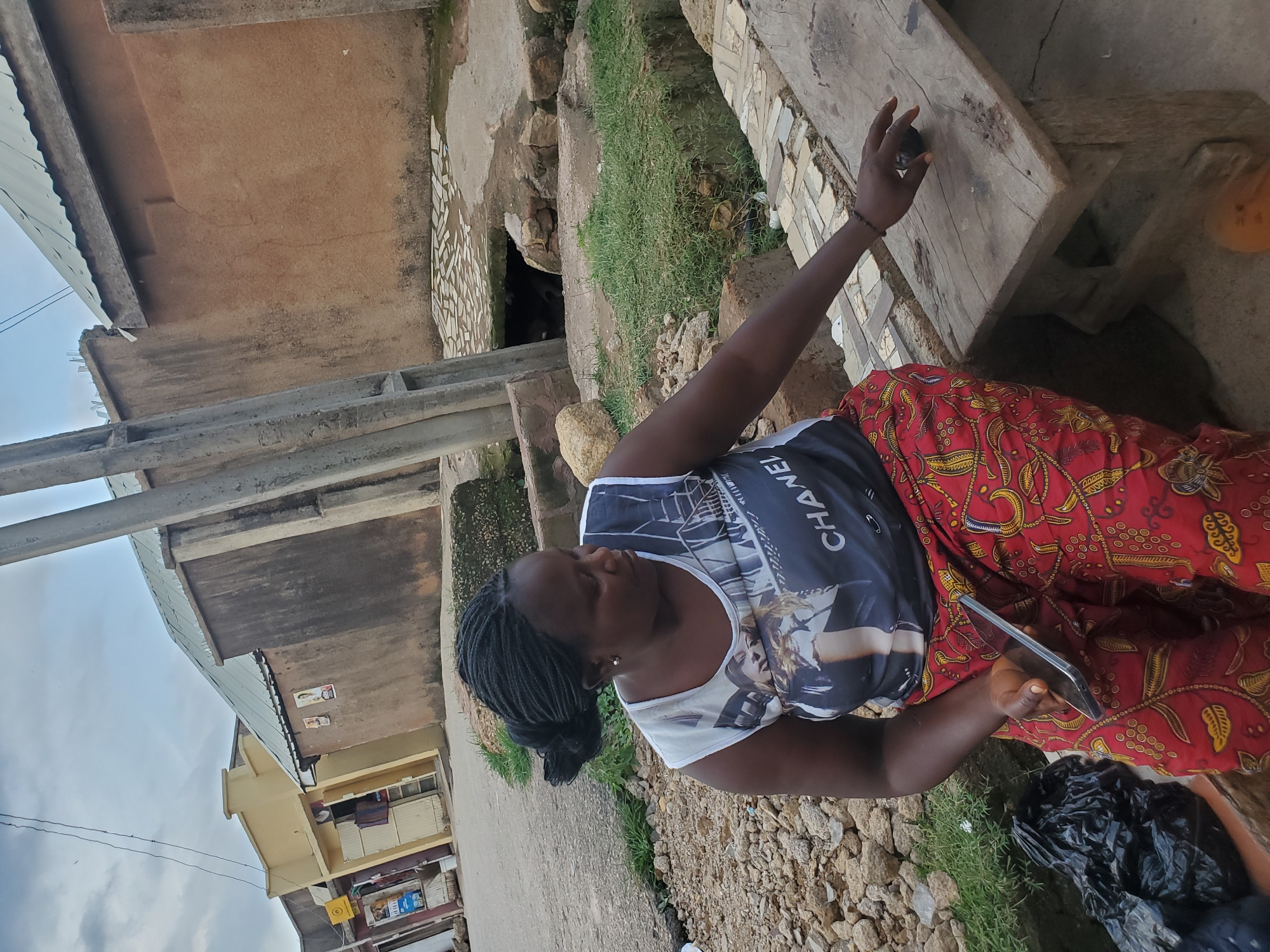 The image shows a woman in an outdoor, semi-urban setting, likely in Jos, Nigeria. She is wearing a white and black t-shirt with "CHANEL" written across the front and a red skirt with a yellow patterned design. Her hair is braided and pulled back. She is holding a smartphone in her right hand and looking down at it. Her left hand is resting on a wooden surface, possibly a table or bench.
The background reveals a mixture of rough terrain, grass, and stone structures, along with some buildings. One building appears to be made of concrete with a corrugated metal roof. Another building in the distance has yellow and beige painted walls with windows and some clothing hanging out to dry. The ground is covered with stones and dirt.
The lighting suggests it is daytime, with the sun casting shadows. The overall atmosphere seems casual and everyday. There are no explicit indicators of a specific event, but the scene depicts a moment of personal engagement with a device in a rustic, possibly residential or market, environment.