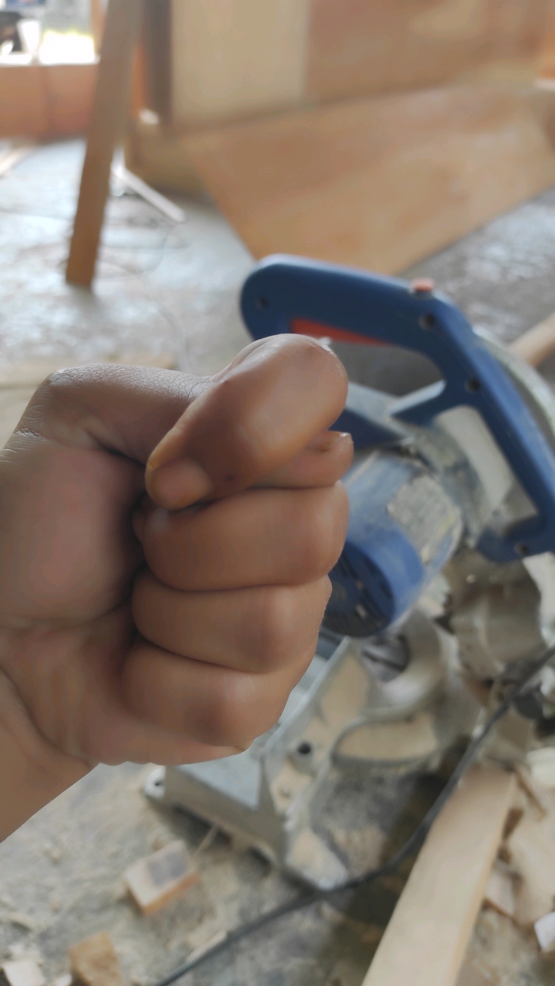 A close-up, vertical, medium shot captures a clenched fist in the foreground, obscuring a portion of a compound miter saw in the background. The hand, possibly female based on its shape and size, is light-skinned and positioned to the left of the frame, with fingers curled tightly into a fist. The saw, predominantly blue and gray, appears to be on a workbench or floor, surrounded by sawdust and wood scraps, suggesting a woodworking environment. The lighting is dim, with a warm undertone, and the focus is shallow, drawing attention to the fist. The scene is likely indoors, possibly a workshop, with indistinct elements of a building visible in the background, including a wooden post and a blurred wall. The setting does not provide specific clues to pinpoint a location within Guwosari, Indonesia.