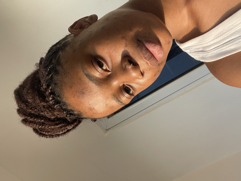 A close-up, slightly angled portrait of a person with dark skin and braided hair. The face is the focal point, revealing a nose ring and an ear piercing. A white tank top peeks into the frame on the right. Sunlight highlights the skin. The background is a combination of a window frame and a white ceiling. The overall composition creates a casual and intimate feel.
