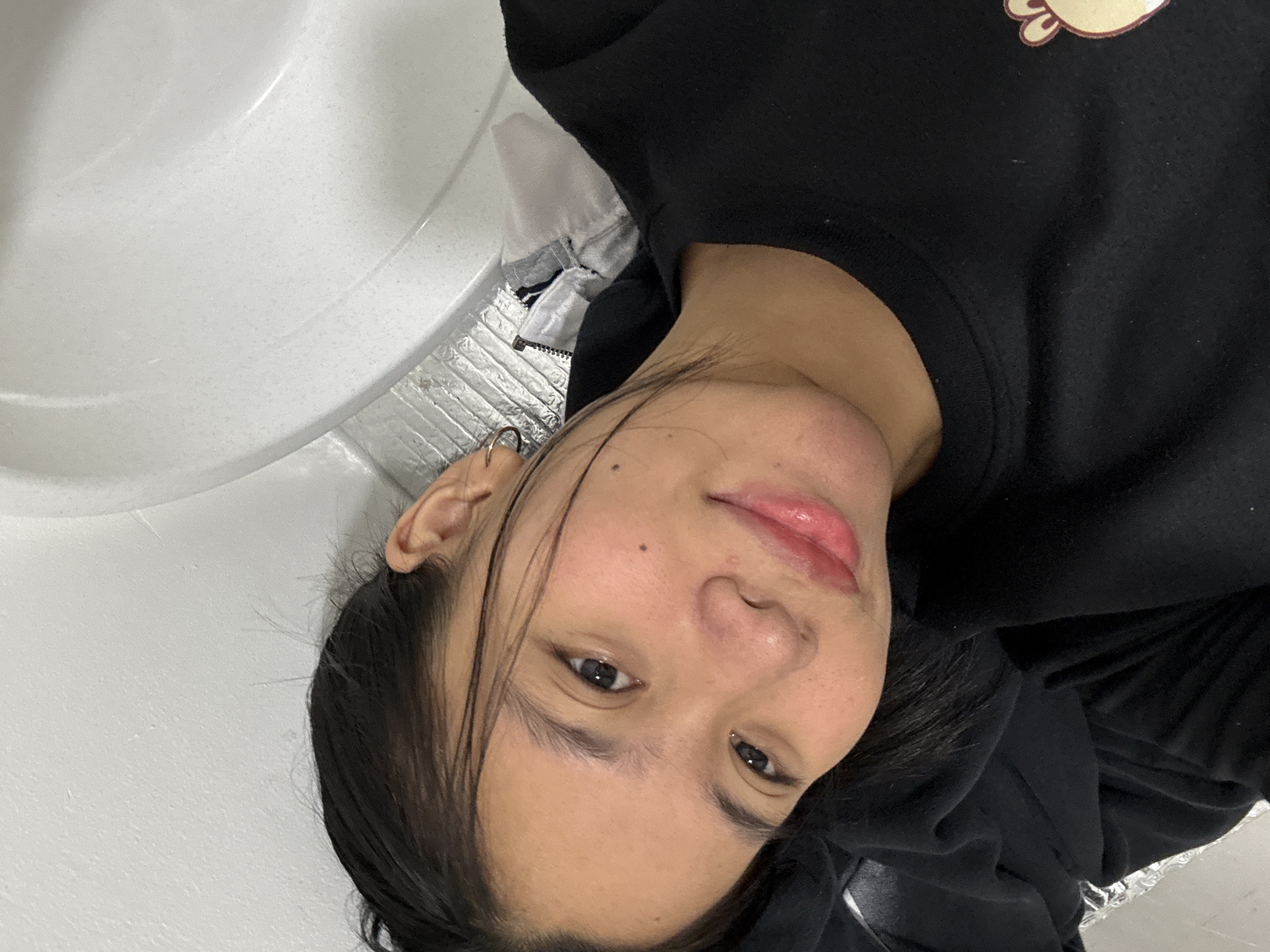 A close-up, high-angle selfie shows a young East Asian woman with dark hair pulled back. She is wearing a black crew neck shirt with a cartoon animal graphic on the right side of the chest. The woman is lying down, with her head resting on a black surface, possibly clothing. Behind her is a white, curved surface that resembles the inside of a bathtub or sink. To the left of the white surface is a grey and white zippered jacket. The background appears to be a bathroom. The image was taken indoors, likely in South Jakarta, Indonesia, based on the inferred context.