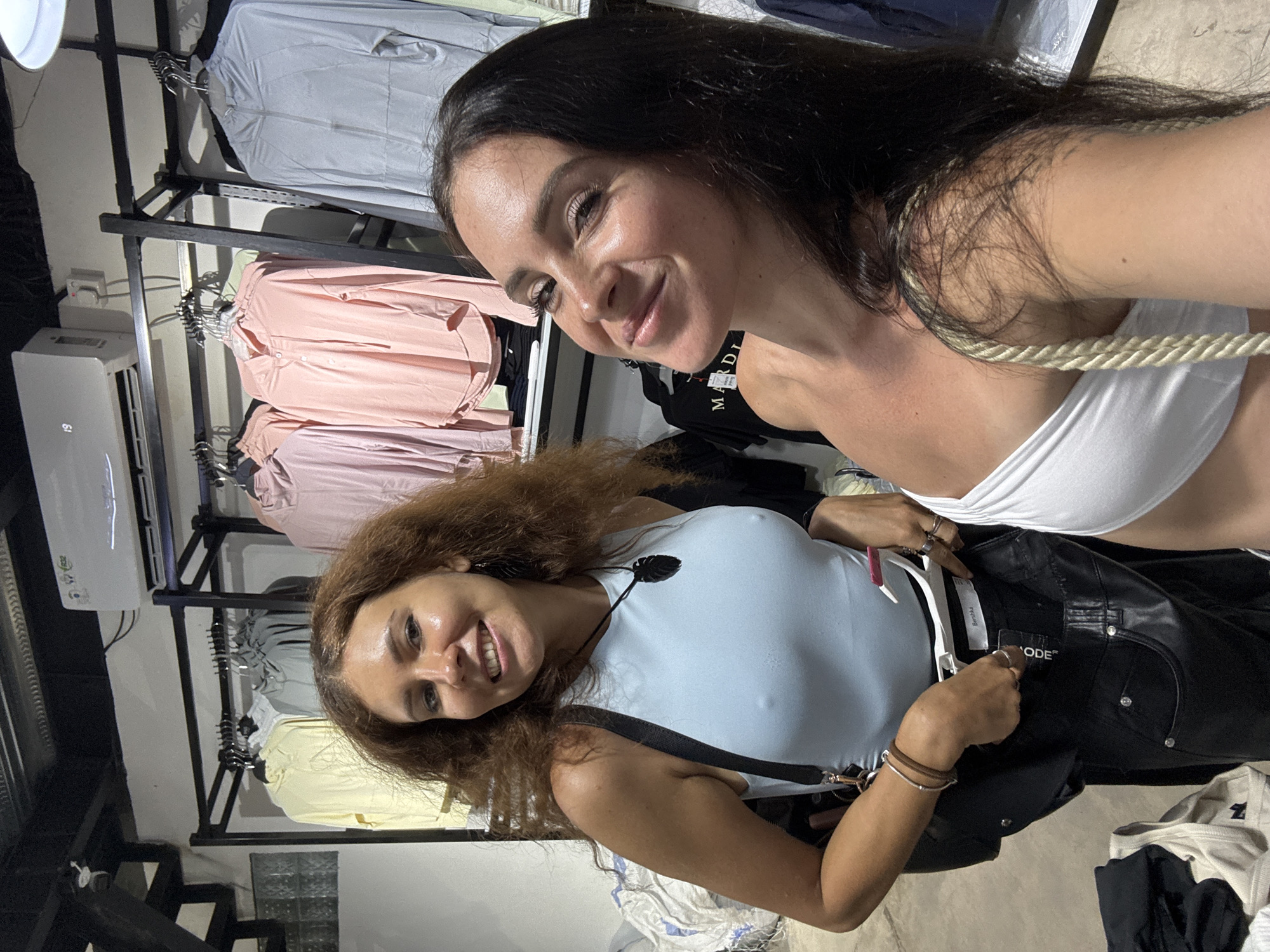 Two women are taking a selfie in a clothing store, possibly located in North Kuta, Indonesia. The woman on the left has long, curly brown hair and is wearing a light blue sleeveless top and black pants. She is holding up a pair of black pants and a clothes hanger. A black bag is slung over her shoulder, and she is wearing a dark pendant necklace and a bracelet. The woman on the right has long, dark hair and is wearing a white strapless top. She is smiling at the camera. Behind them, clothing racks are filled with various garments, including pastel-colored hoodies and grey jackets. An air conditioning unit is mounted on the wall above the clothing racks. The store appears to be well-lit, with a modern aesthetic.